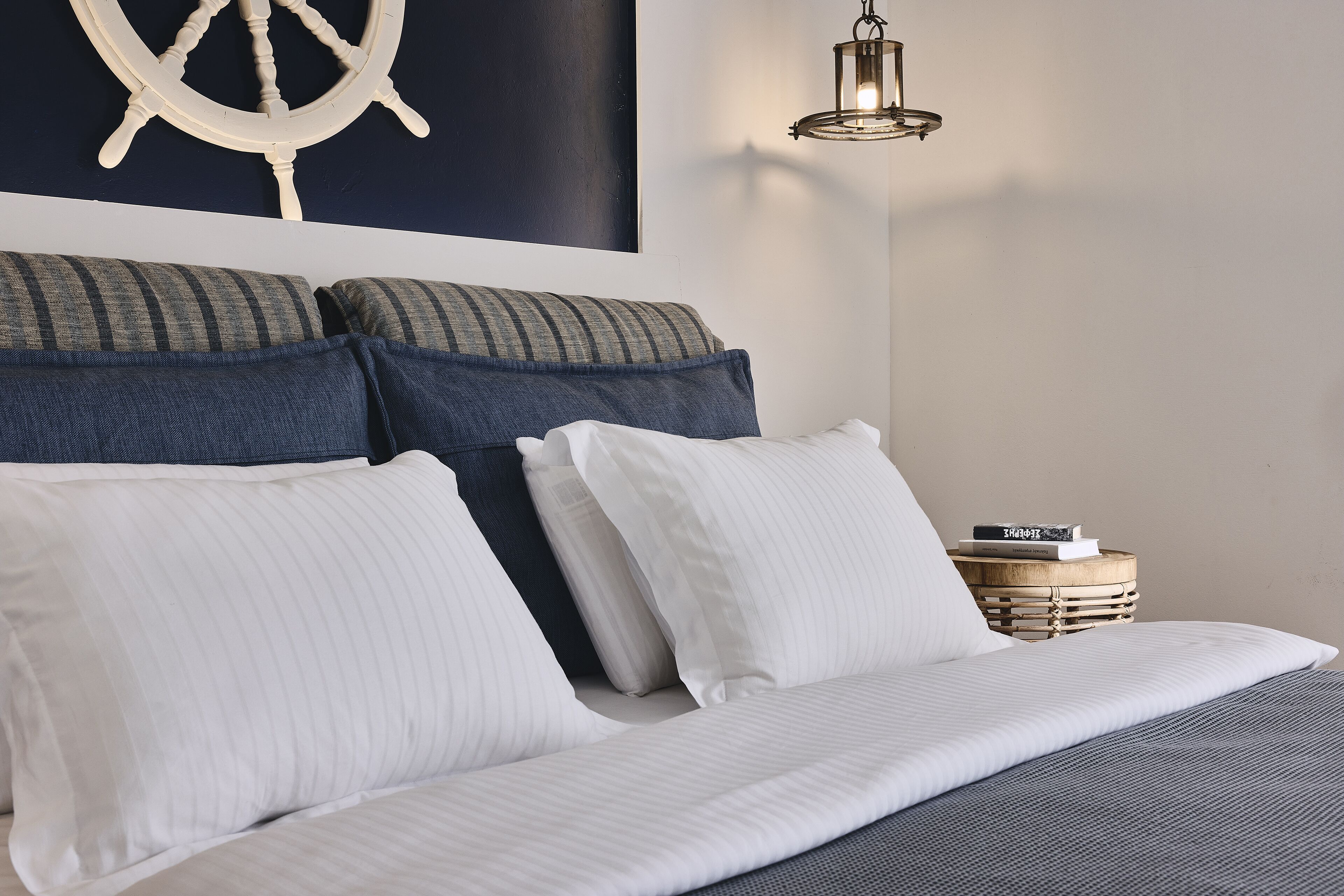 Una cama bien hecha con ropa de cama blanca y a rayas, un cabecero con varios cojines, un adorno con timón de barco en la pared y una mesita de noche con un farol.