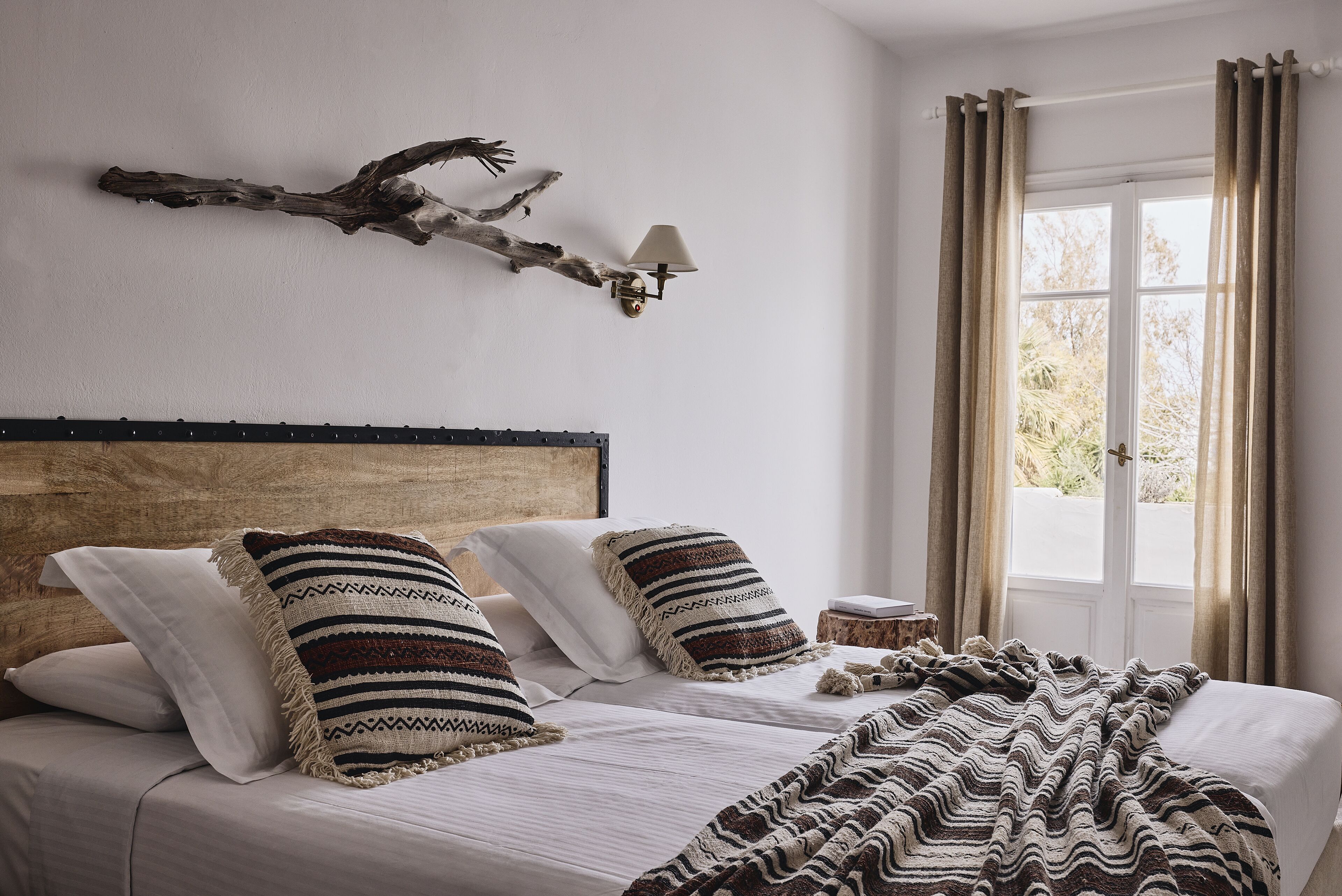 A bedroom with a wooden headboard, two beds with striped pillows, a branch wall art piece, a bedside lamp, and a window with curtains.