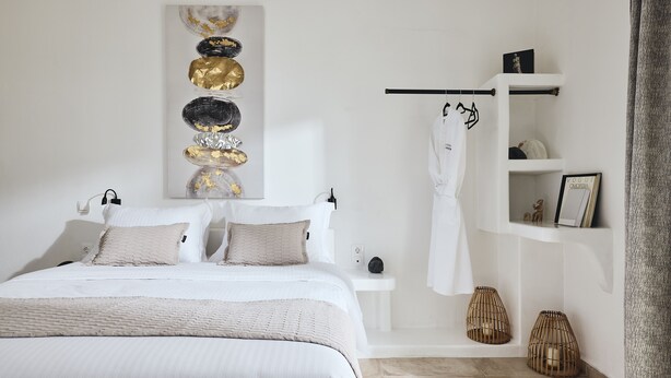 A neatly arranged bedroom with a bed, bedside tables, a robe hanging, and decorative items. - Capetan Giorgantas (Milos)