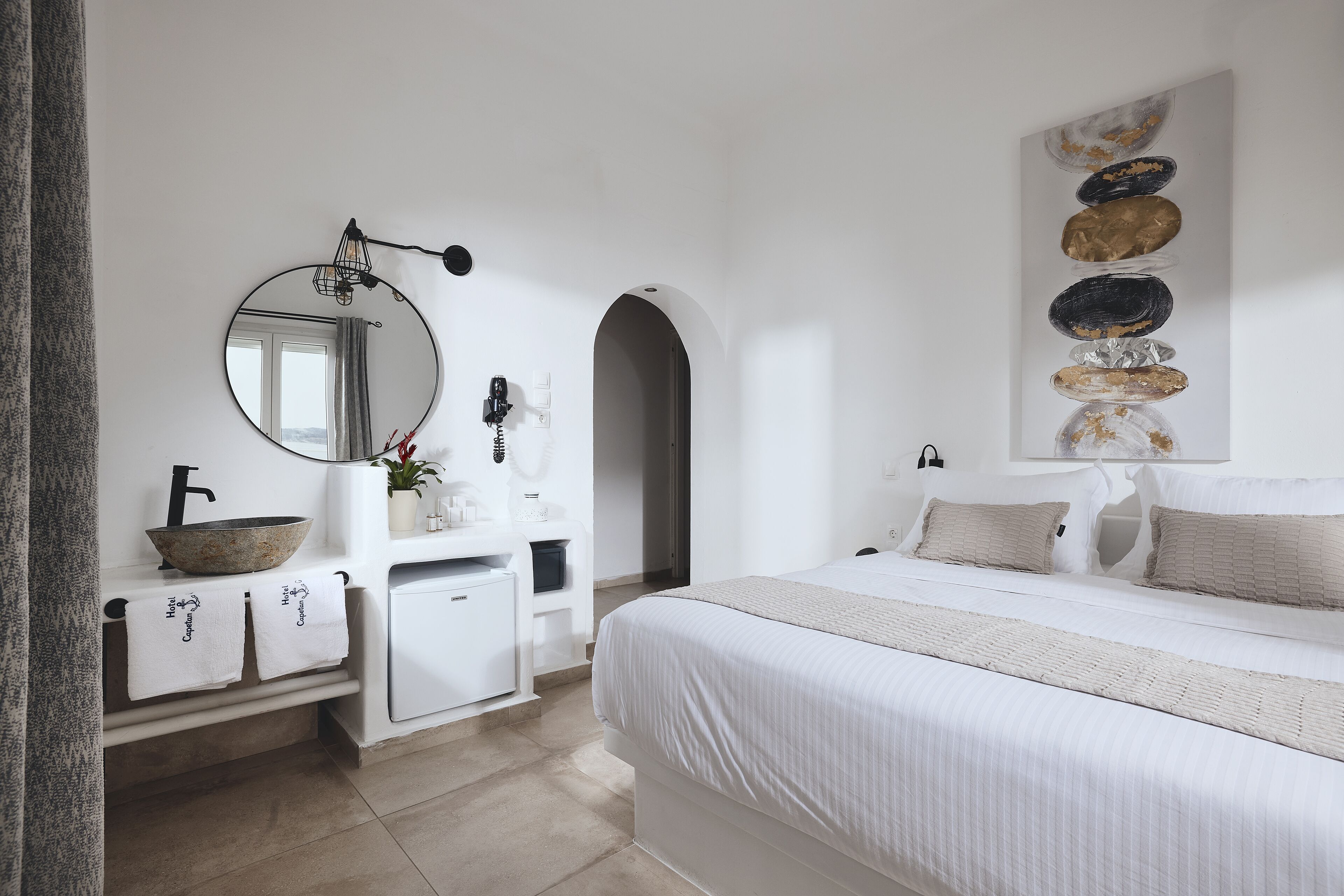 A modern bedroom with a large bed, a round mirror, a white dresser, and a wall-mounted artwork.