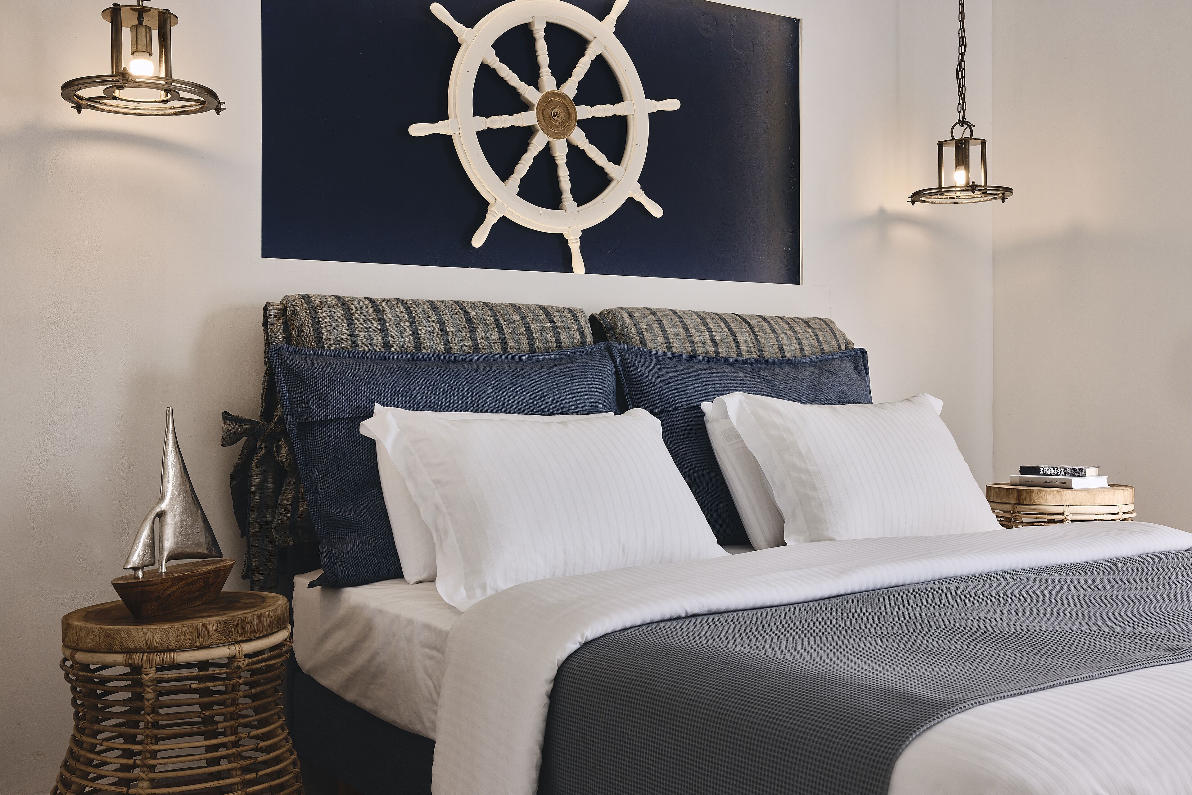 Una cama bien hecha con un edredón a rayas, un timón de barco montado en la pared y dos lámparas colgantes.