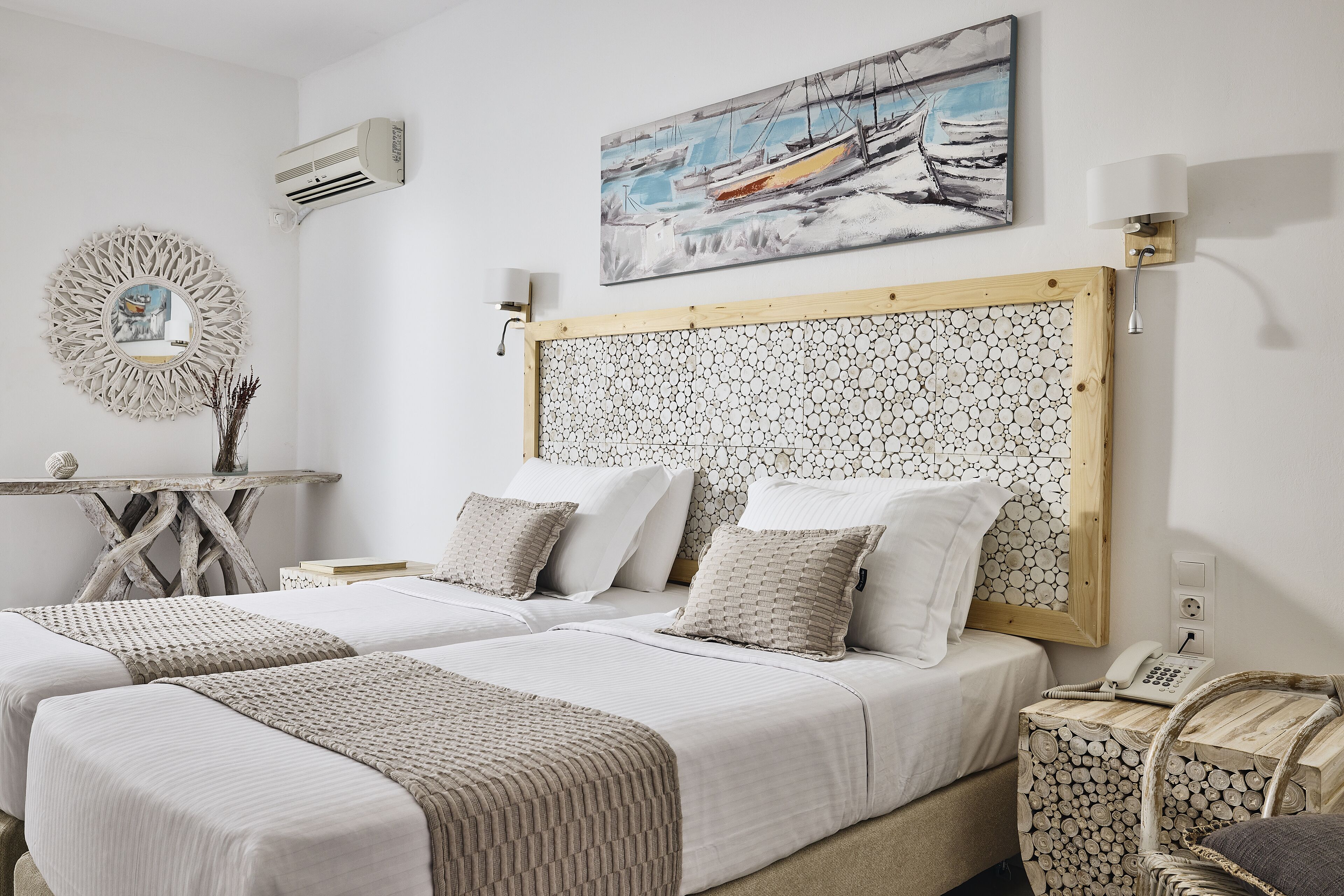 Un dormitorio con dos camas, cabecero de madera, aire acondicionado de pared, un cuadro, un espejo redondo y una mesita de noche con lámpara.