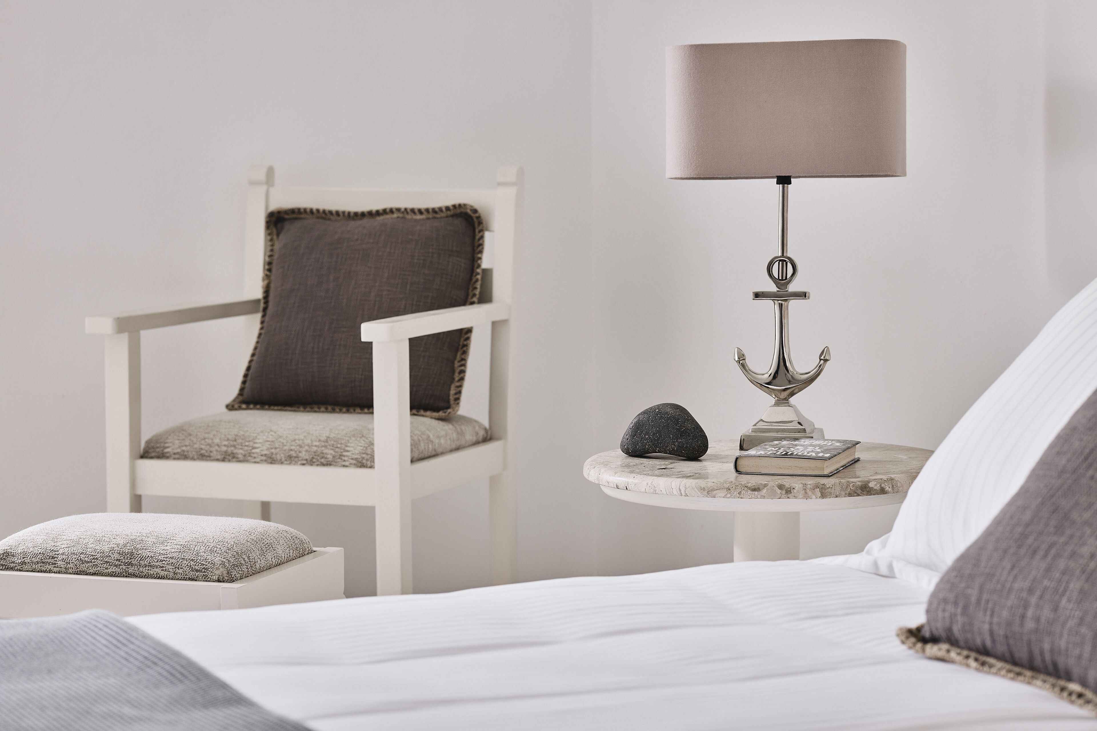 A neatly arranged bedroom with a bed, a white chair with a cushion, a small round table with a lamp, a book, and a decorative item.