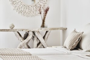 Una cama con edredón blanco y almohadas, una mesita redonda con un florero de plantas secas, y un espejo montado en la pared.