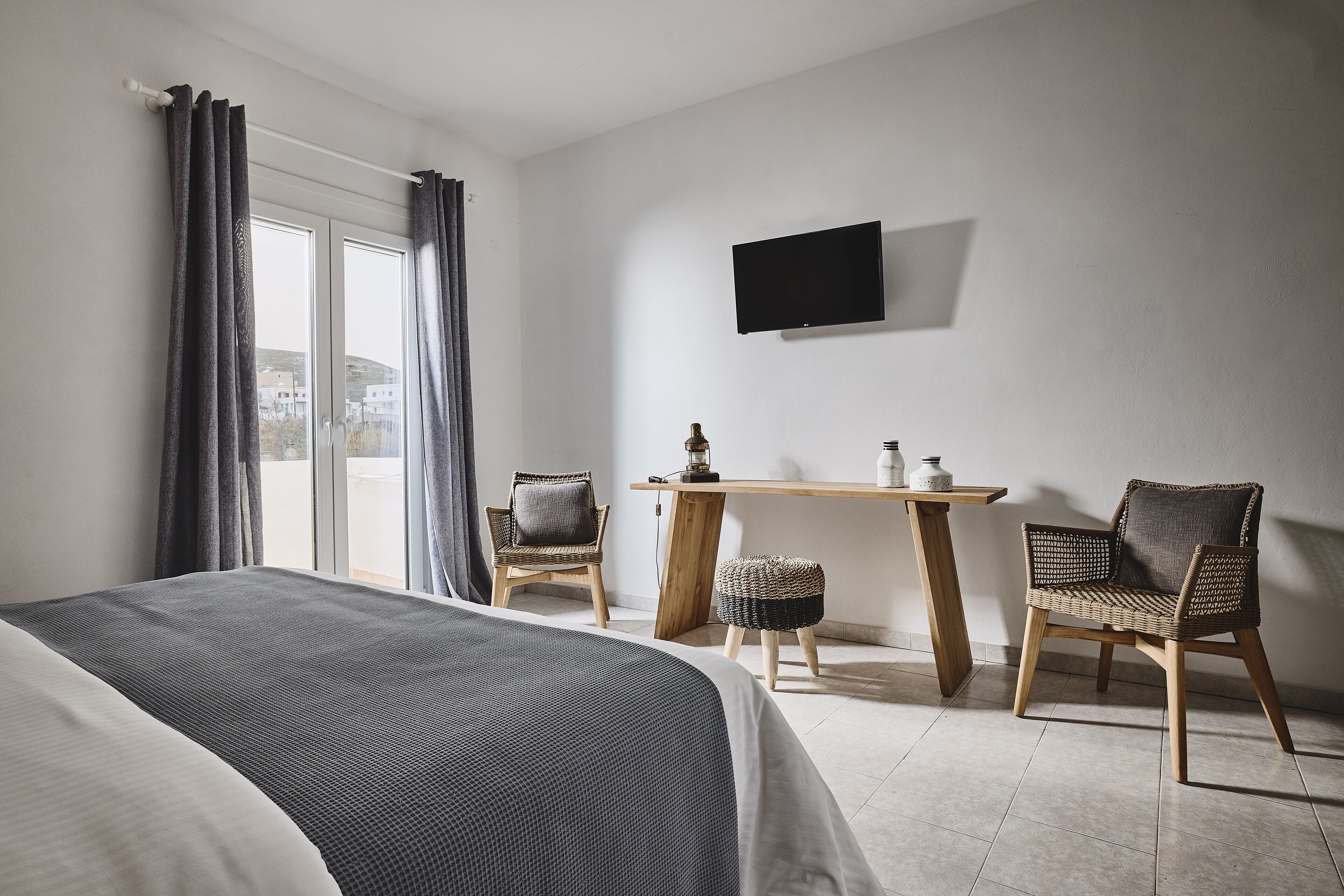 Habitación de hotel con una cama, dos sillas de mimbre, una mesita de madera, un televisor montado en la pared y un ventanal grande con cortinas.