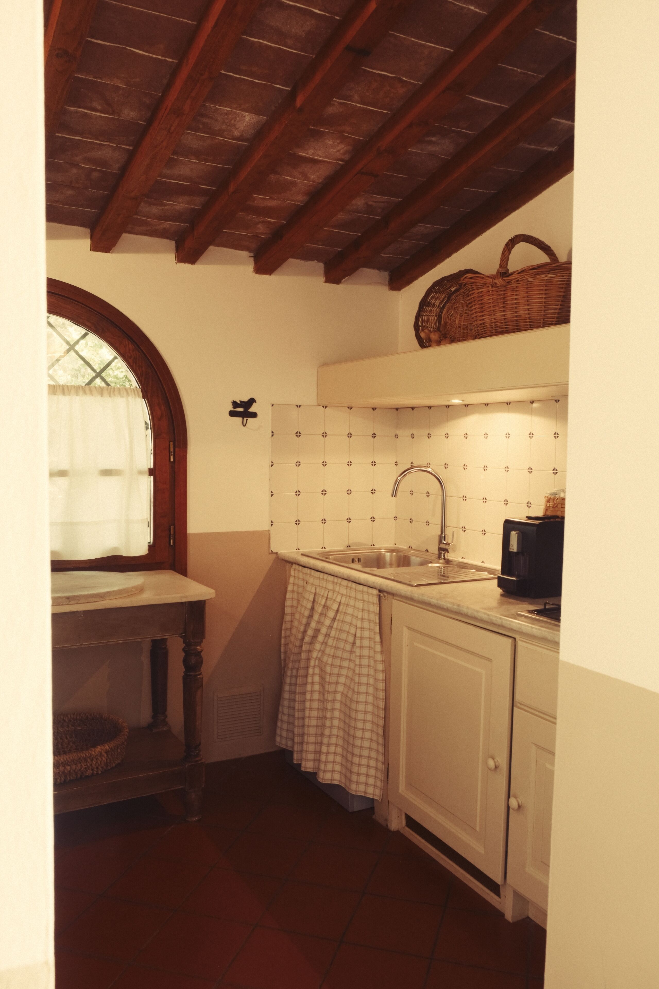 A kitchen with a wooden ceiling, a window with a curtain, a sink, a toaster, and a basket on the wall.