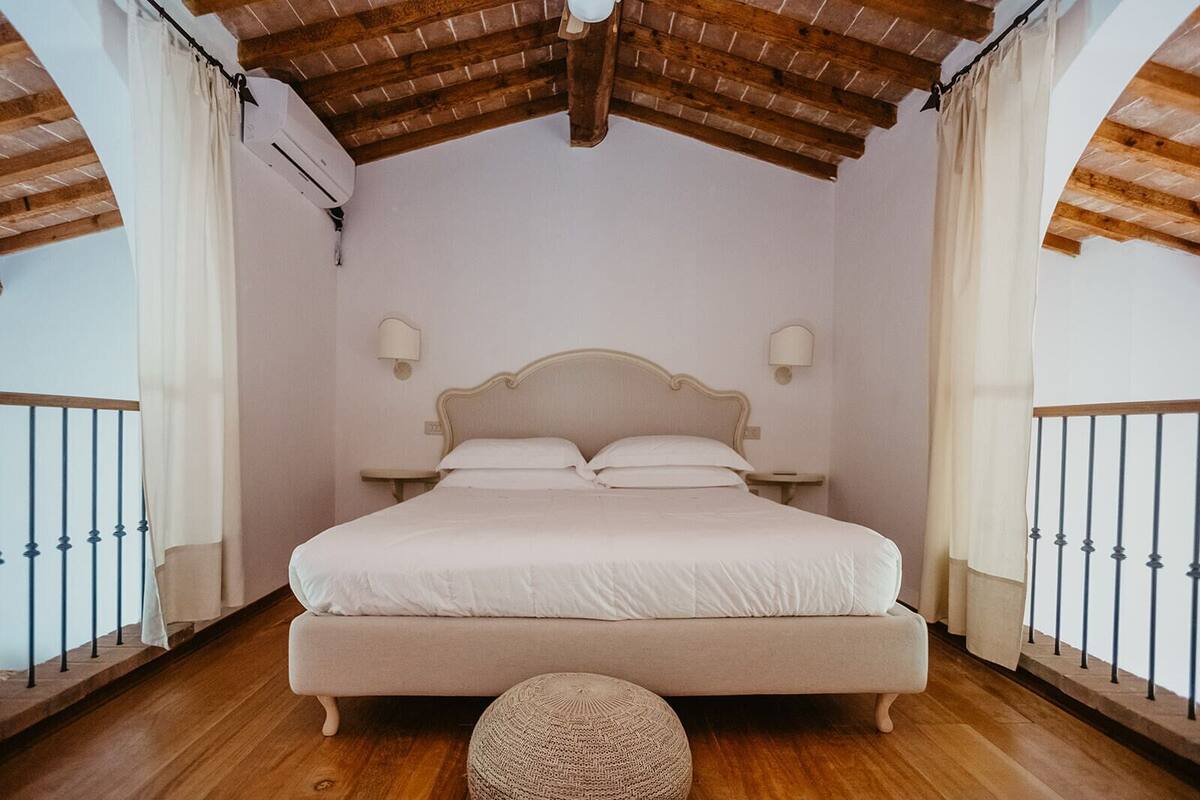 A bedroom with a wooden ceiling, a bed with white bedding, two bedside tables, and a woven ottoman.