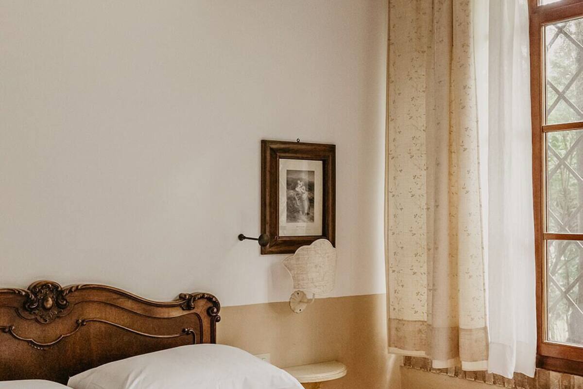 A bedroom with a wooden headboard, a bed with white bedding, a small bedside table, a framed picture on the wall, and a window with curtains.