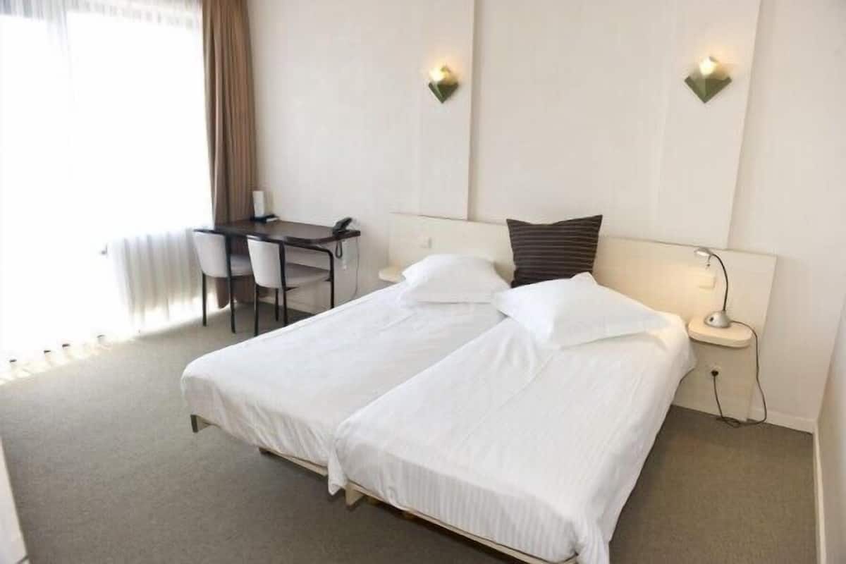 A hotel room with a large bed, a desk, two chairs, and a nightstand with a lamp.