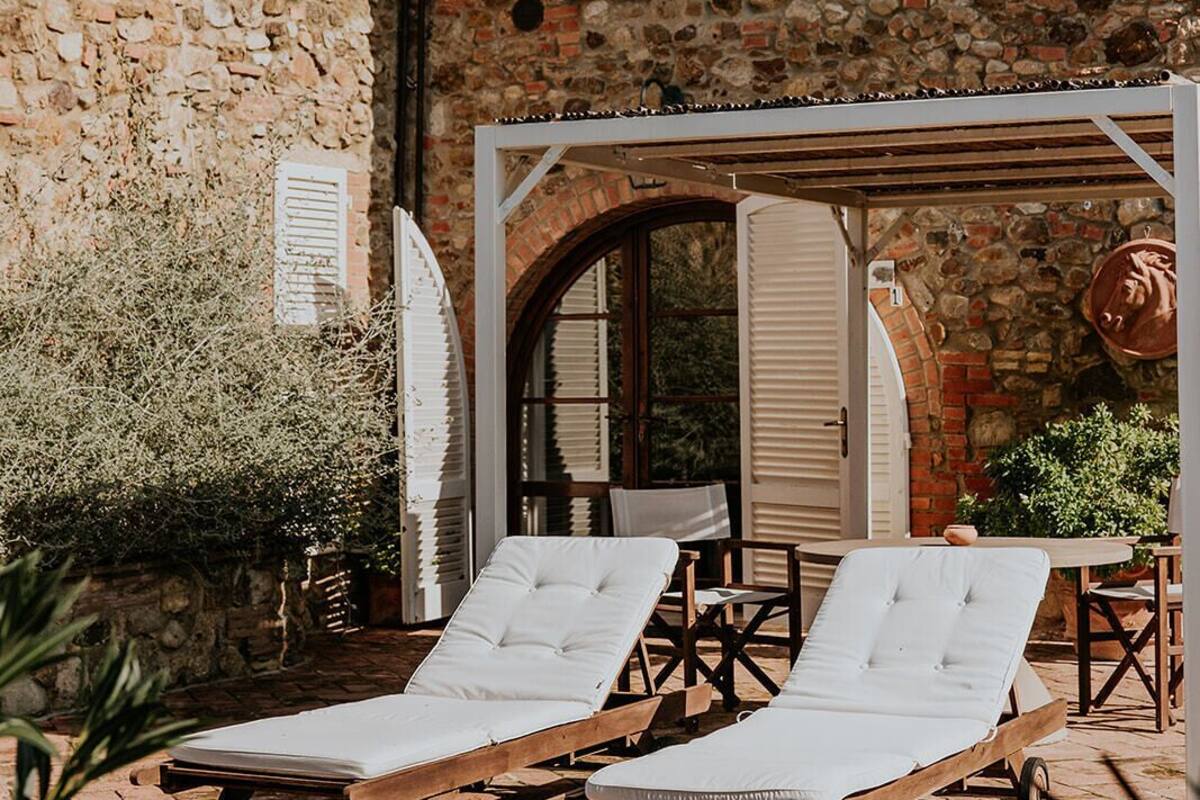 Un bâtiment en pierre avec un toit en briques, une pergola en bois et deux fauteuils de jardin.
