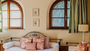A cozy room with a bed, a sofa, and a table. There are two windows with curtains, and a lamp on the table. - Poggio Cornetto (Bibbona)