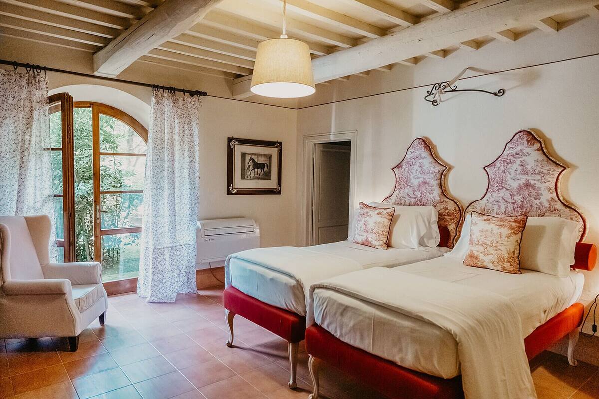 Une chambre avec deux lits, une chaise, une fenêtre avec des rideaux, un tableau au mur et un ventilateur de plafond.