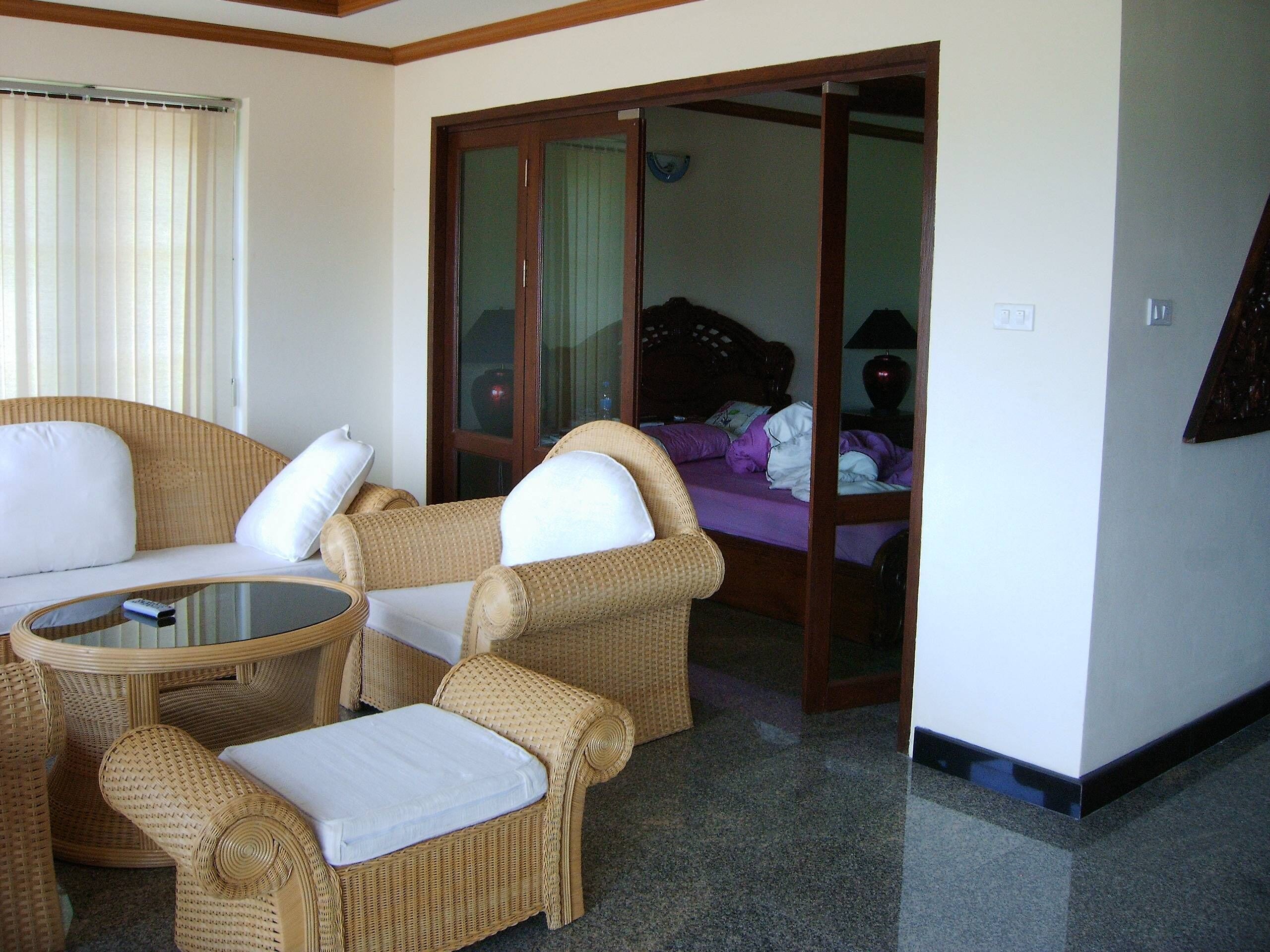 Quarto com móveis de vime, uma mesa de vidro e uma cama com roupa de cama roxa.