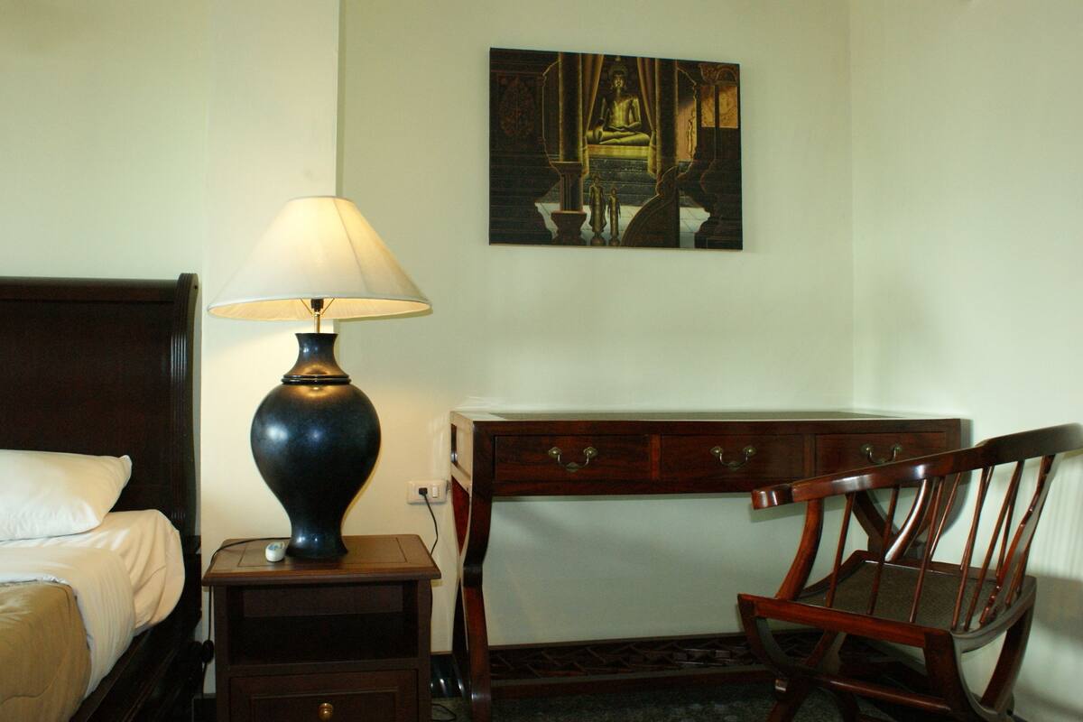 A hotel room with a bed, a nightstand with a lamp, a wooden chair, a desk with drawers, and a painting on the wall.