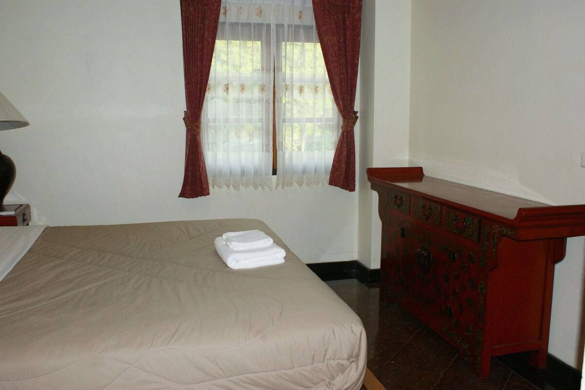 A room with a bed, a wooden dresser, a window with curtains, and a lamp.