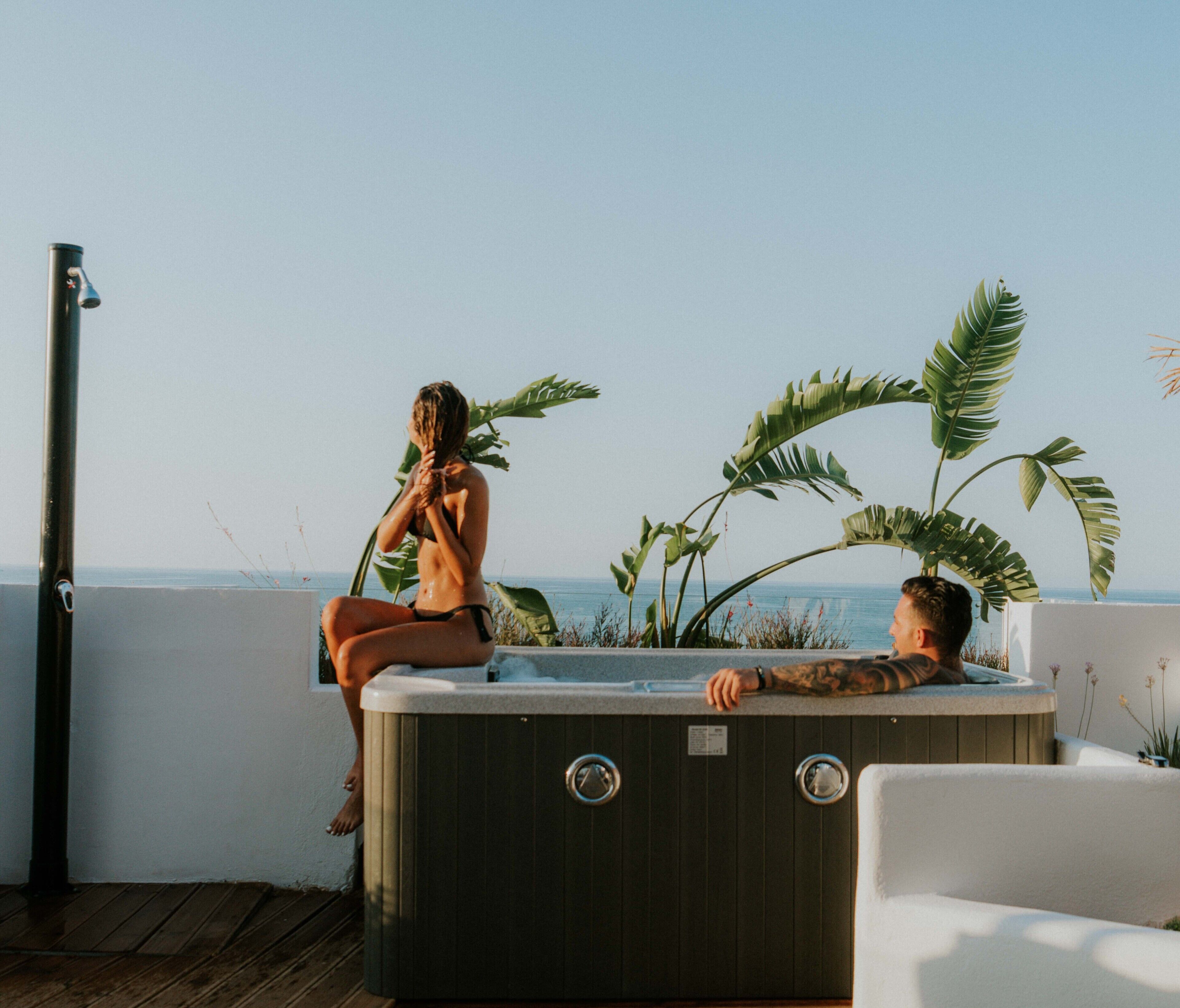 A hot tub with a woman and a man sitting in it, surrounded by tropical plants, overlooking a body of water.