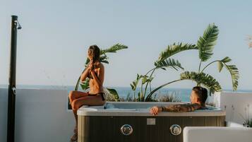 A hot tub with a woman and a man sitting in it, surrounded by tropical plants, overlooking a body of water.