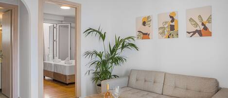 A modern living room with a sofa, a wooden coffee table with a bottle and glass, a large potted plant, and artwork on the wall.