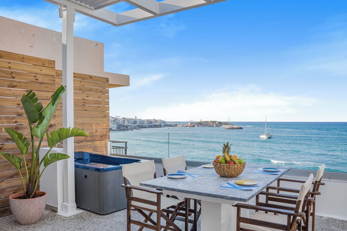 A terrace with a table set for dining, overlooking a coastal view with boats and a cityscape in the distance.