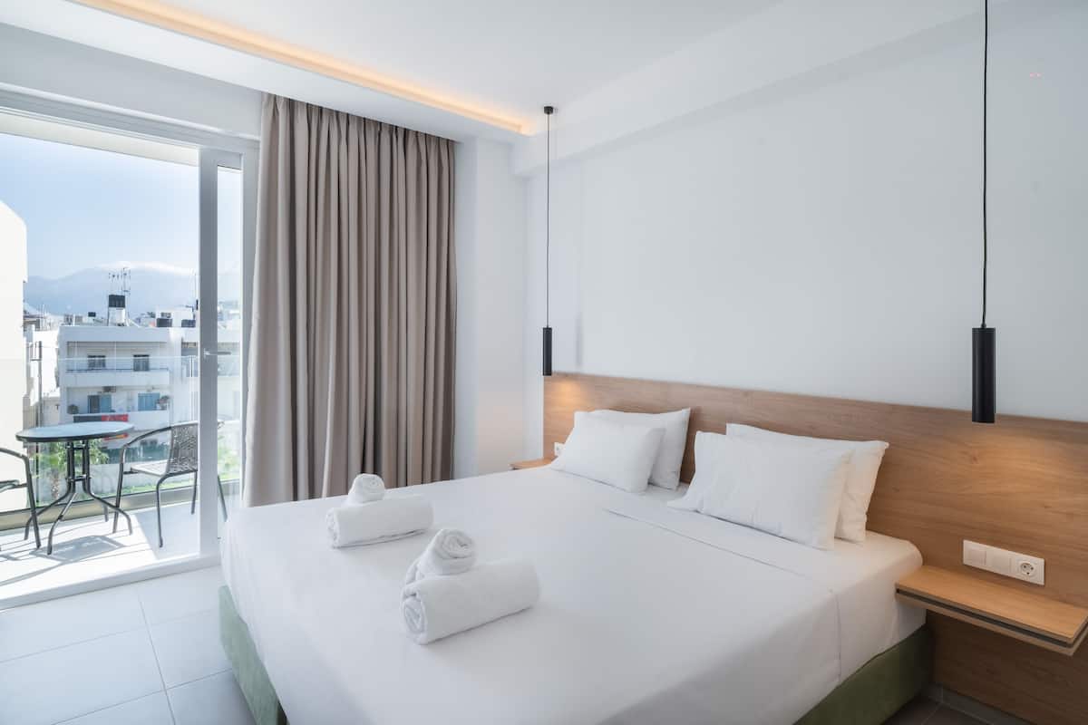 A hotel room with a large bed, a wooden headboard, a balcony with a table and chairs, and a view of buildings and mountains.