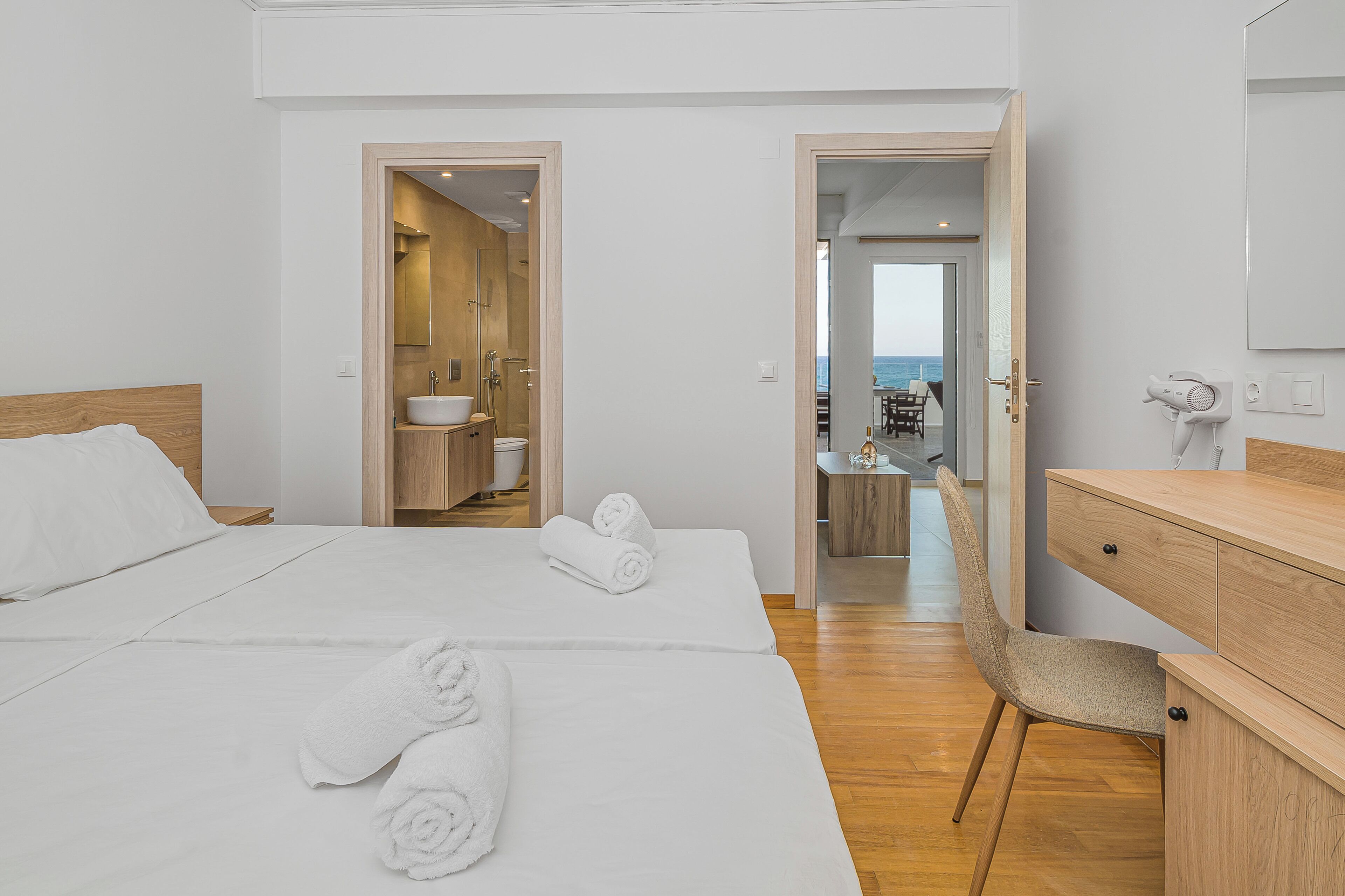 A hotel room with two beds, a wooden desk, a chair, a bathroom, and a view of the sea.
