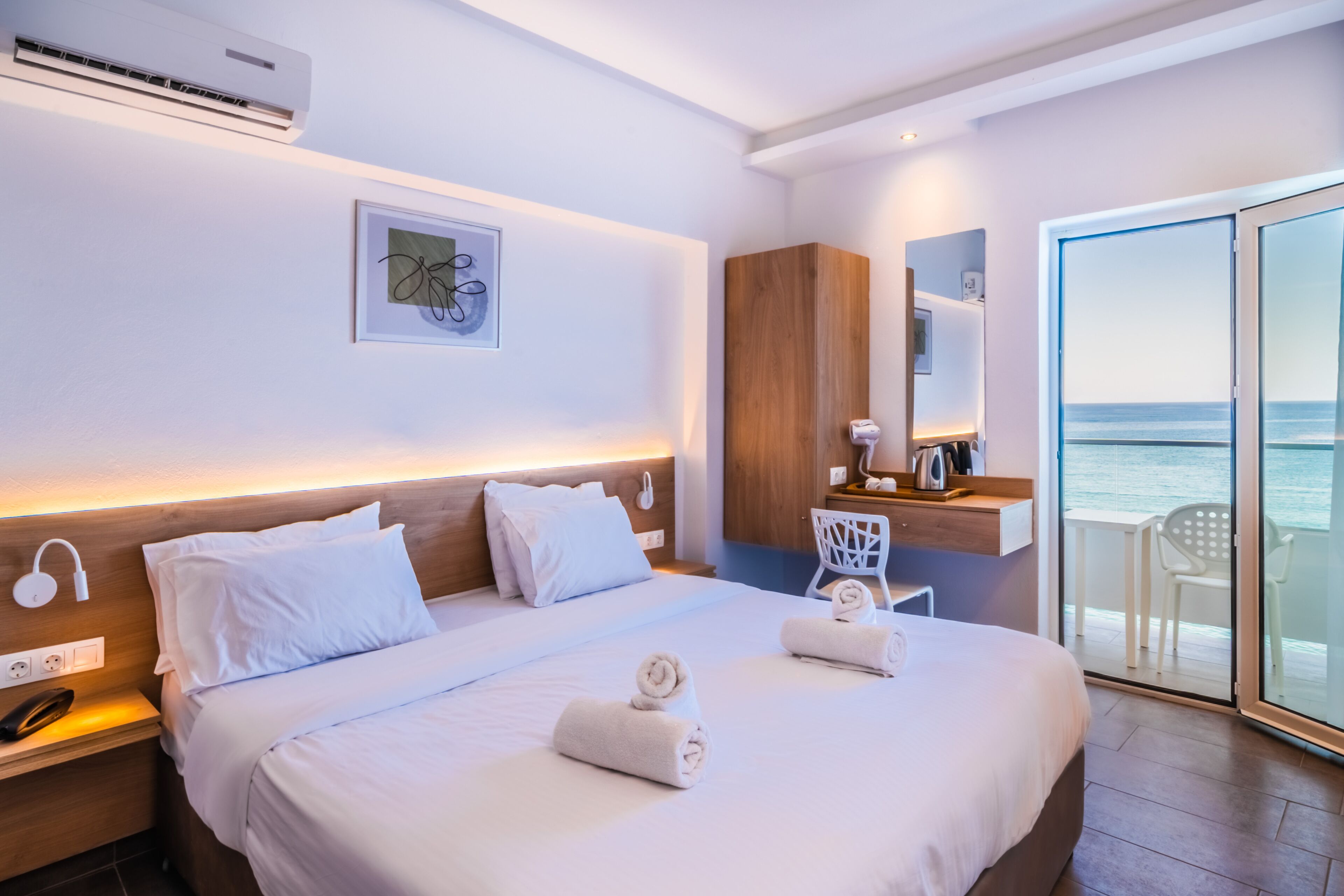 A hotel room with a bed, a bedside table with a phone, an air conditioning unit, a wall-mounted artwork, and a view of the sea through a glass door.