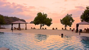 Ein Poolbereich mit Liegestühlen und Sonnenschirmen, im Hintergrund ein Strand und ein Sonnenuntergangshimmel.
