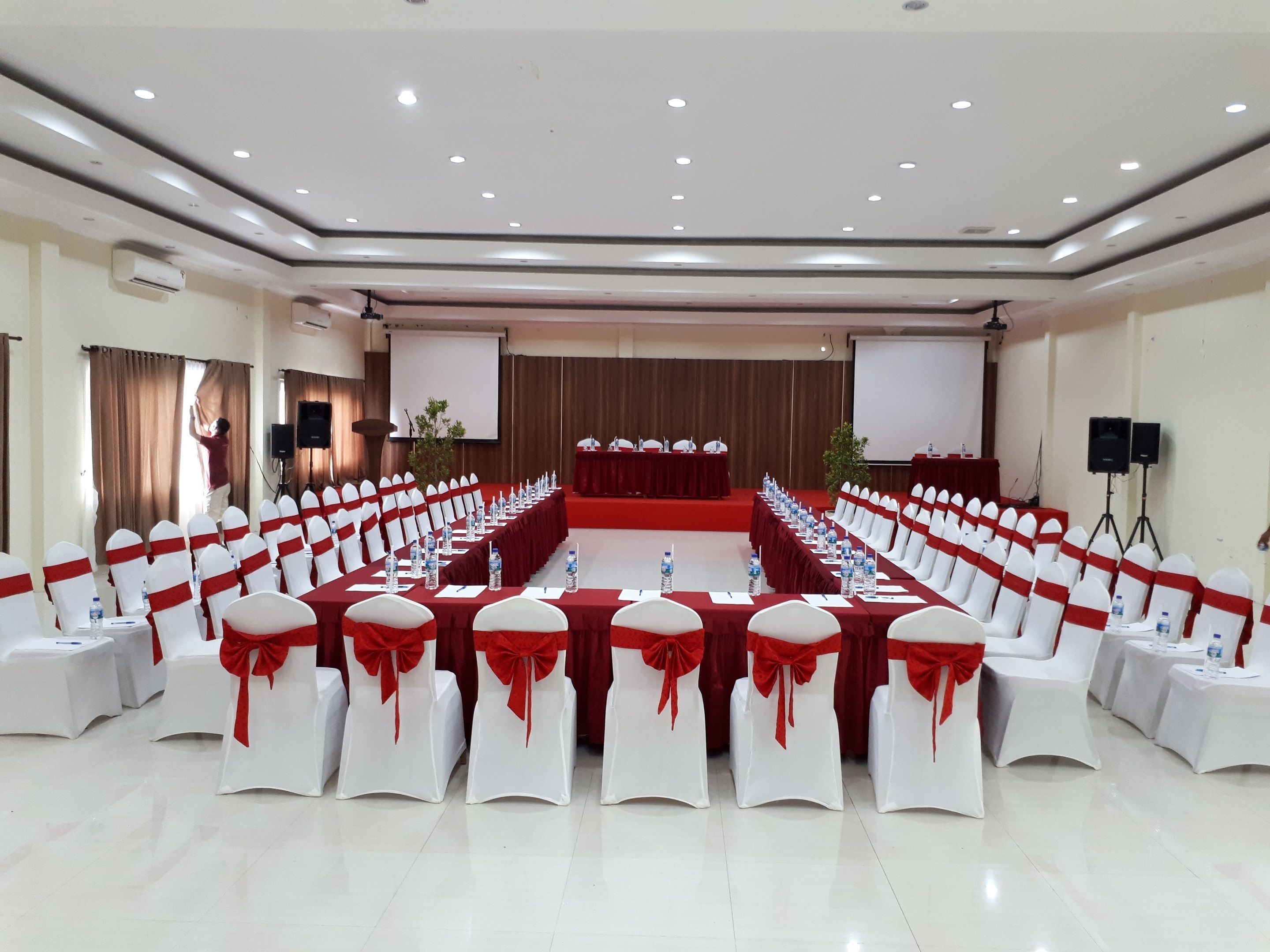 Une salle de conférence avec une longue table, des chaises blanches ornées de rubans rouges, un pupitre et un écran de projection.