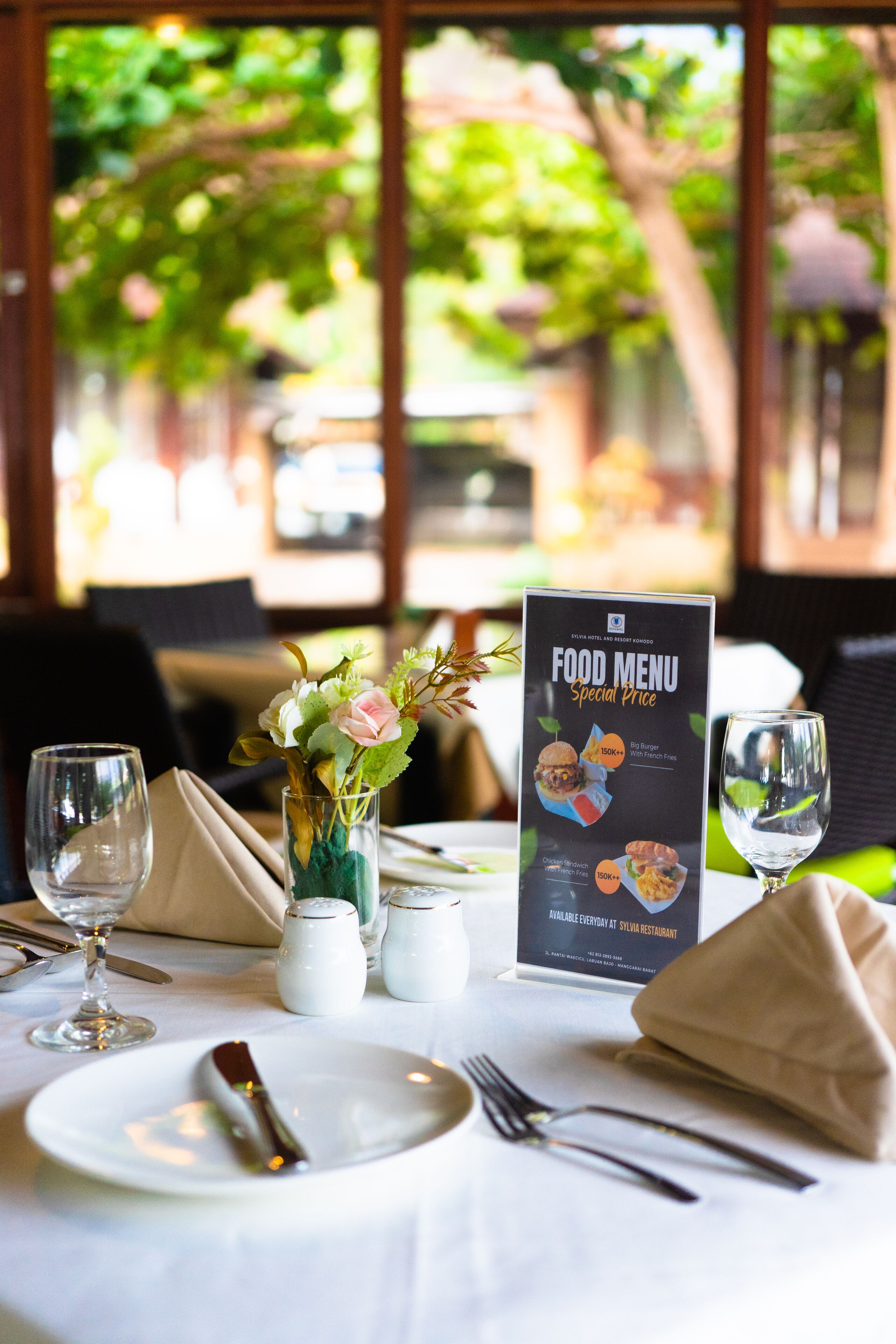 A table set for dining with a food menu, a vase with flowers, and a view of greenery outside.
