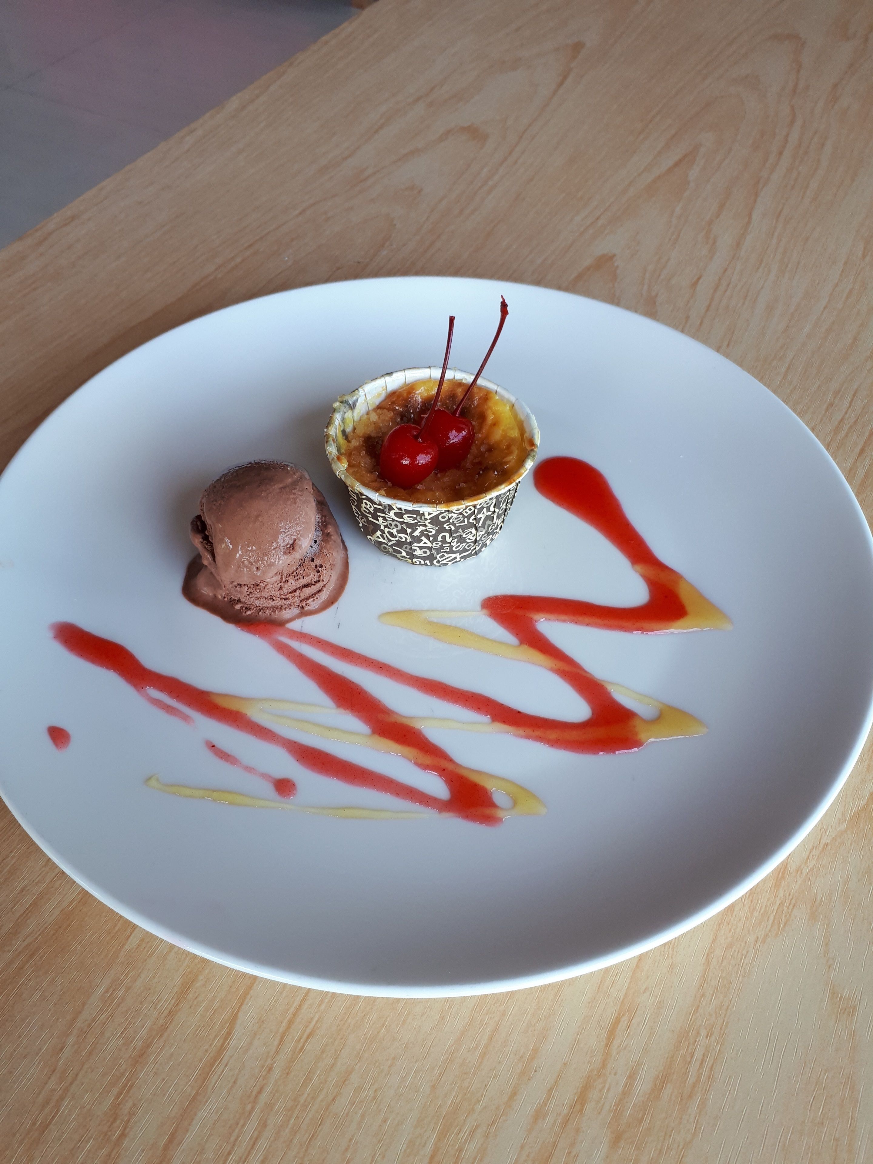 Un plato de postre con helado de chocolate y una tarta con cereza, acompañado de salsas rojas y amarillas.