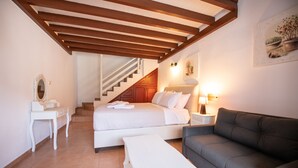 A bedroom with a bed, a nightstand, a bedside table, a sofa, a lamp, and a staircase leading to another room. - Hovolo Hotel Apartments (Skopelos)