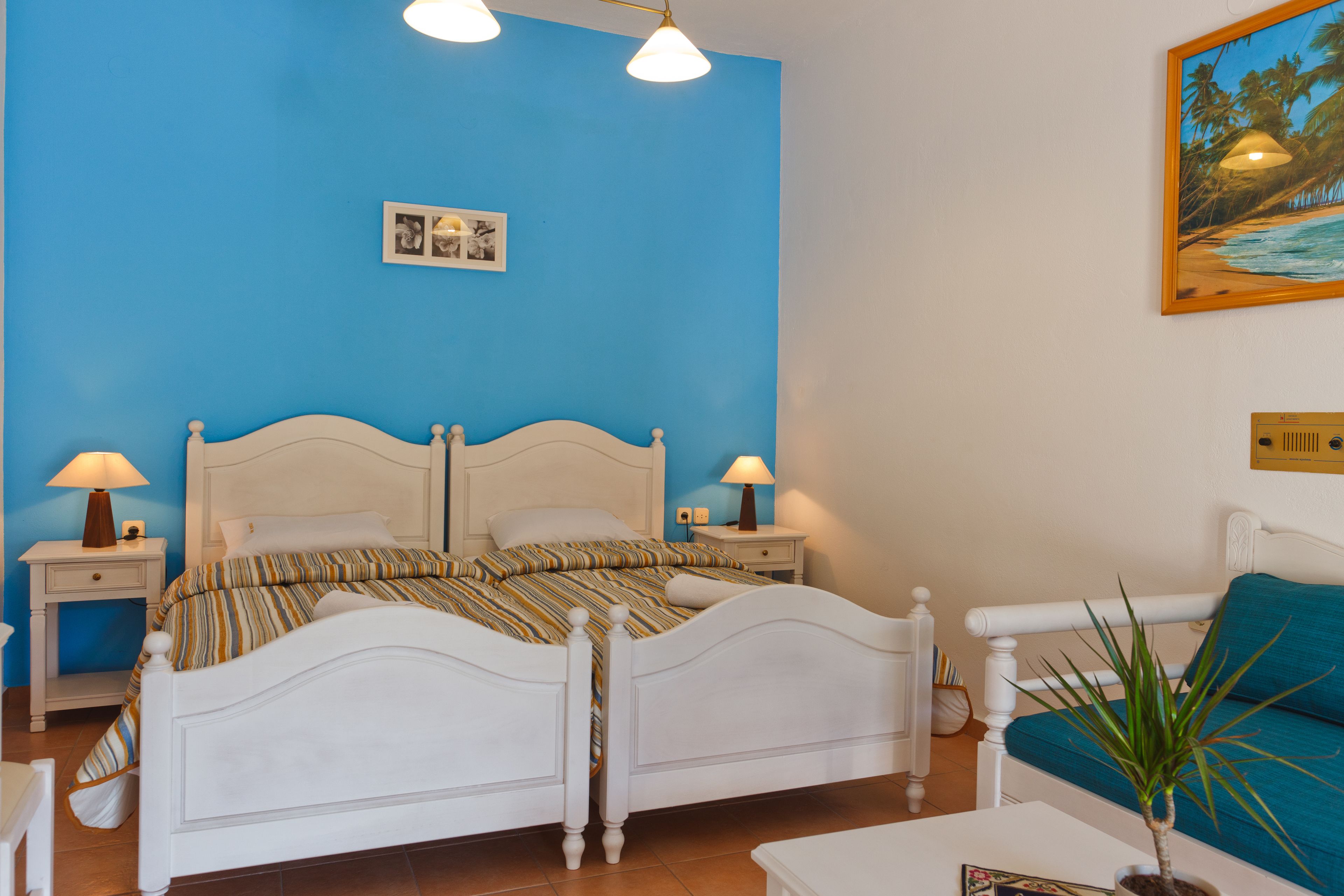 A bedroom with a white bed, two bedside tables with lamps, a blue wall, and a painting of a beach scene.