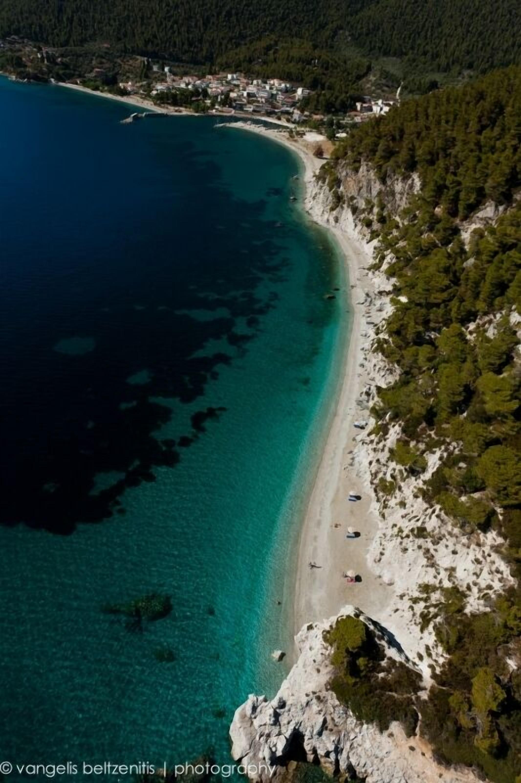 Vue aérienne d’une zone côtière avec une plage, des eaux turquoise et limpides, et une colline boisée.