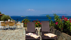 A terrace with wicker chairs and a table, overlooking a coastal view with a stone pathway and potted plants. - Hovolo Hotel Apartments (Skopelos)