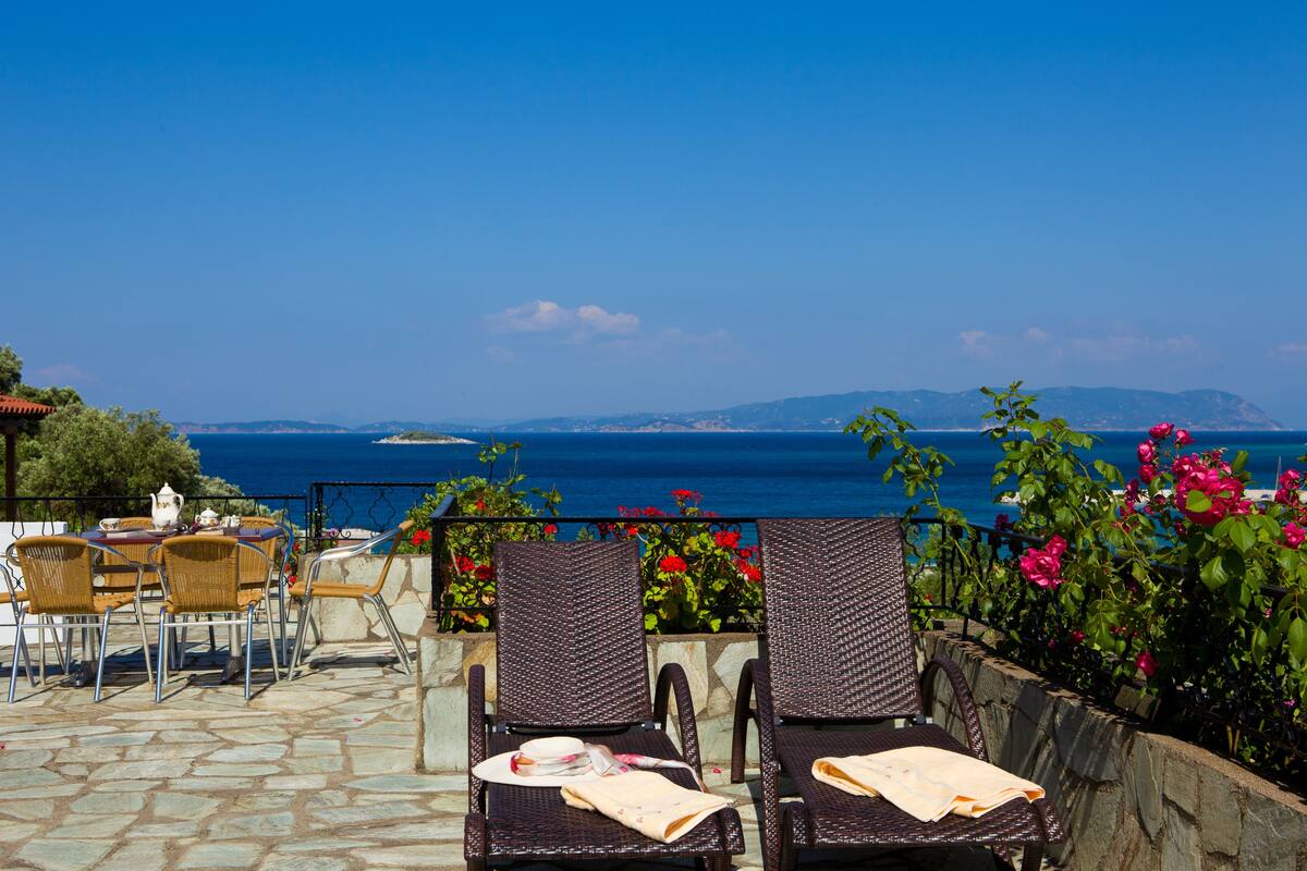 Une terrasse dotée de chaises et d’une table en osier, offrant une vue sur la côte, avec un sentier en pierre et des plantes en pot.