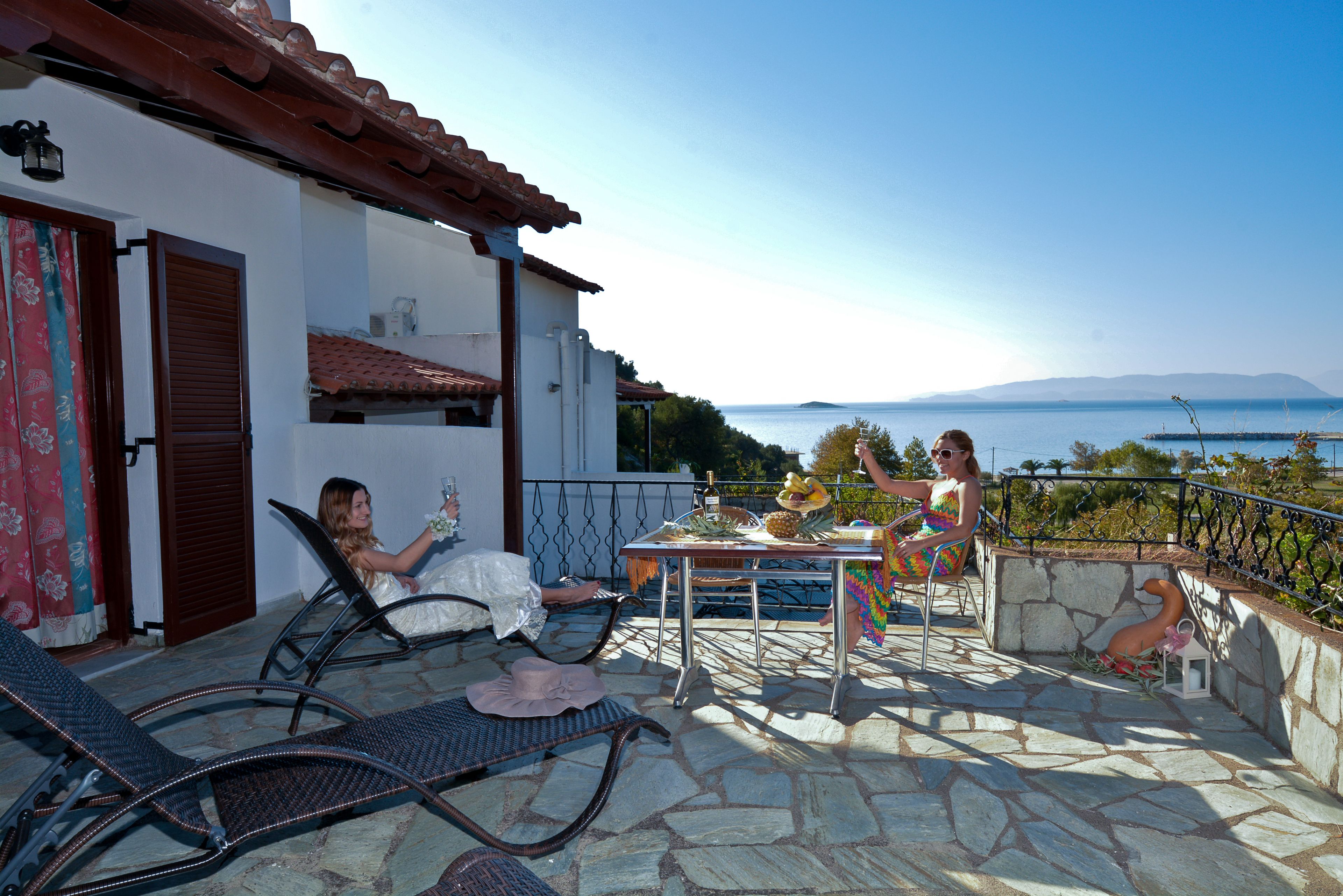 A terrace with stone flooring, a table with fruits and drinks, and two people seated.