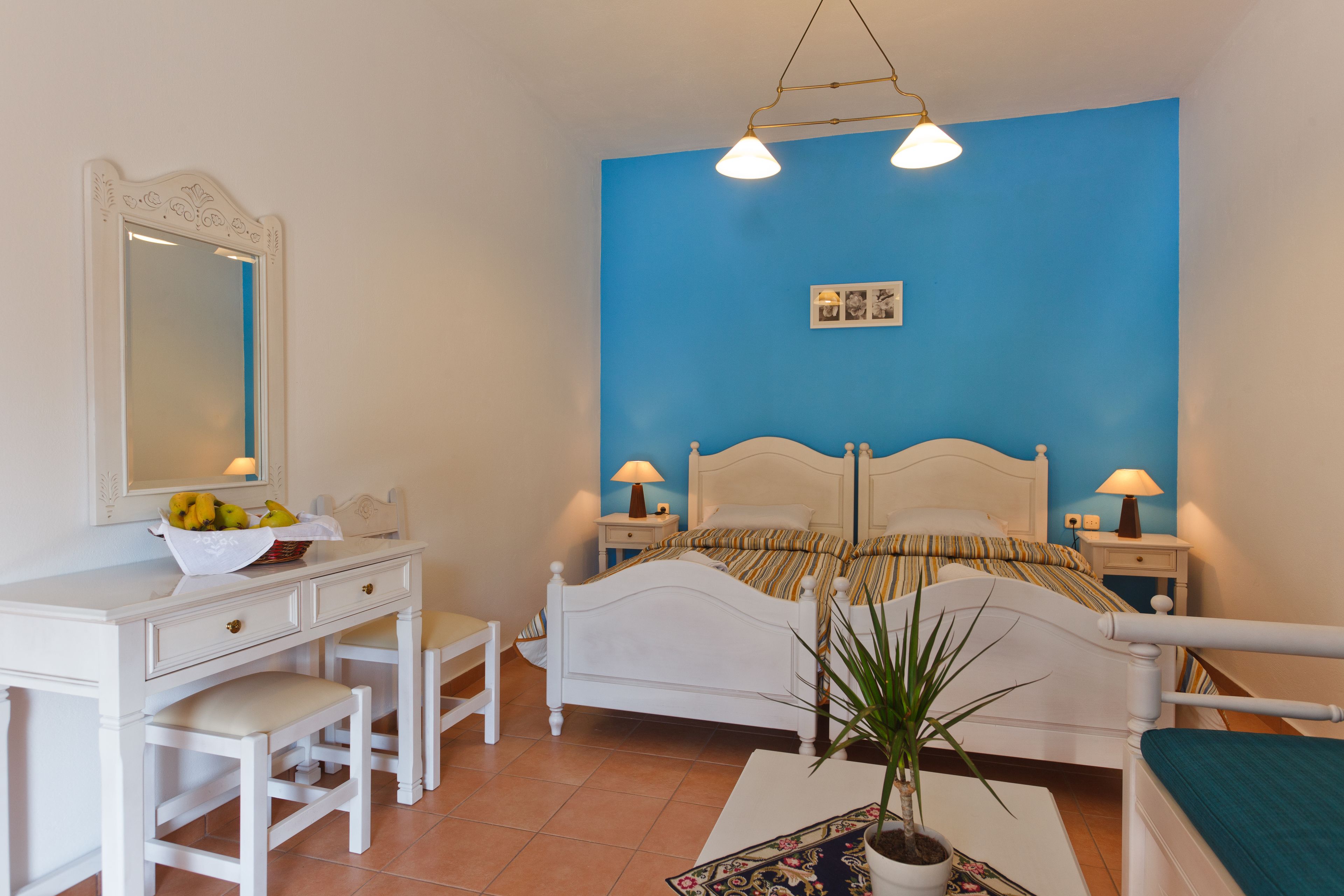 Un dormitorio con una cama blanca, dos mesitas de noche con lámparas, un aparador blanco con espejo y una planta en maceta.