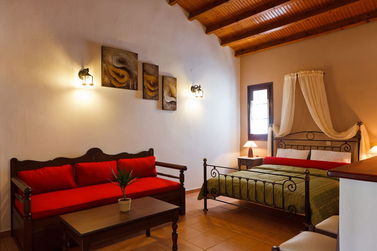 Une chambre avec un plafond en bois, un lit recouvert d’une couverture verte, un canapé rouge, une petite table avec une plante et des œuvres d’art accrochées au mur.