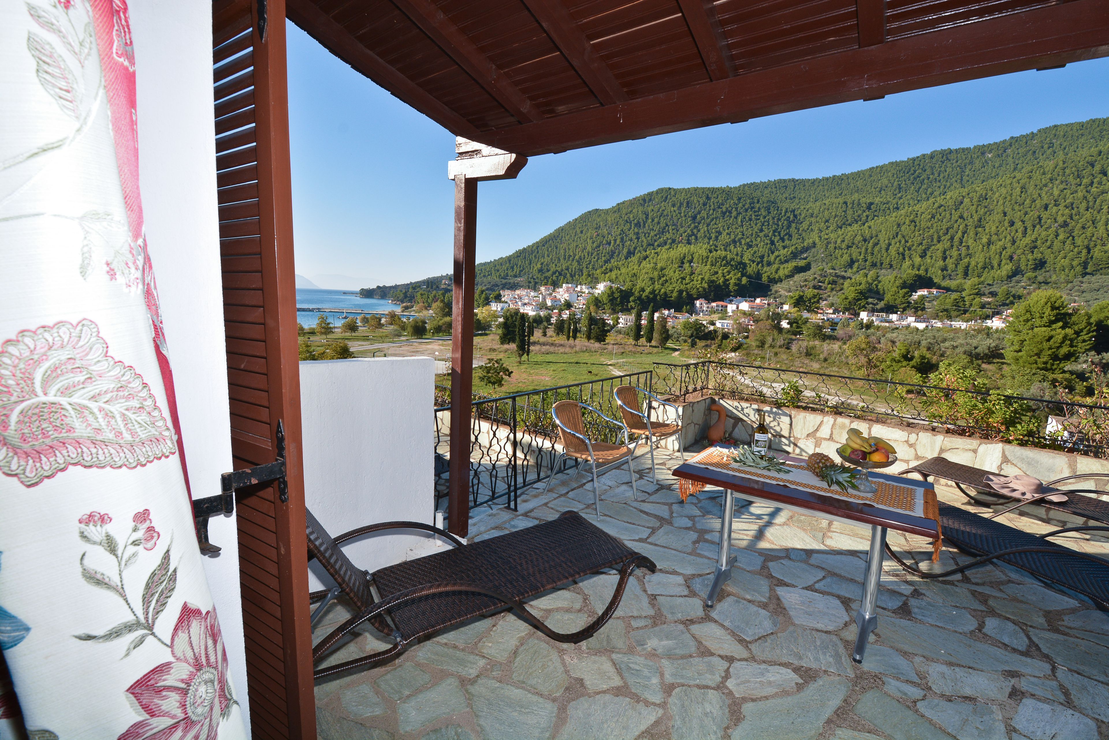 A stone patio with a view of a town and mountains, a lounge chair, and a table with fruit.
