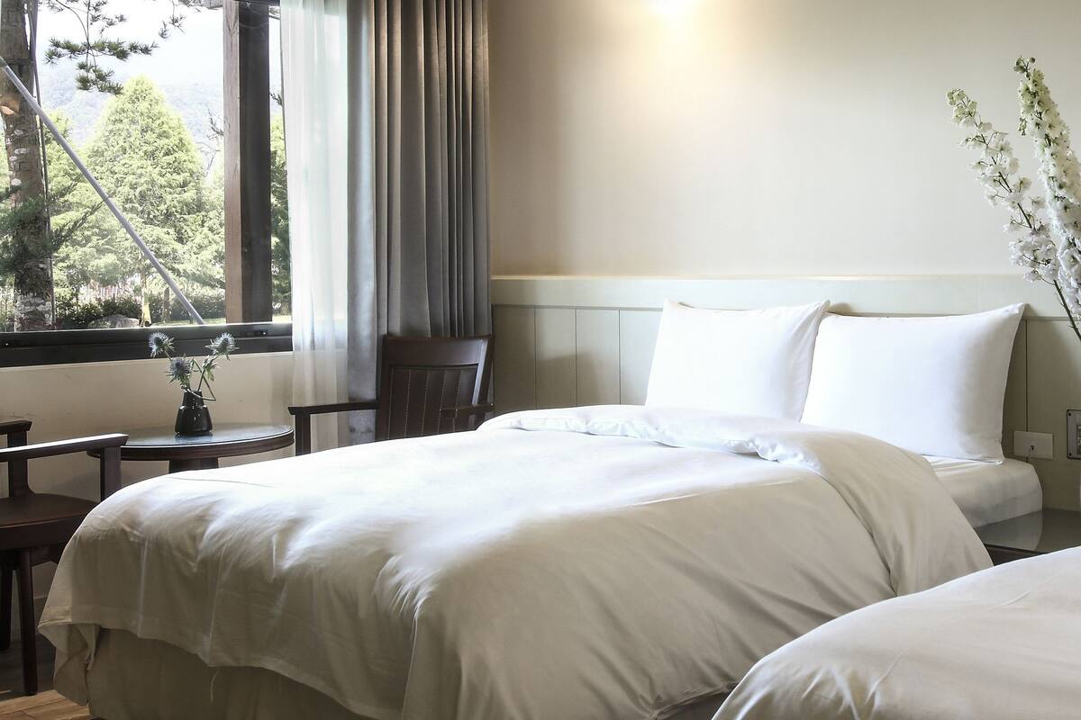 A hotel room with two beds, a small table, and a window with curtains.