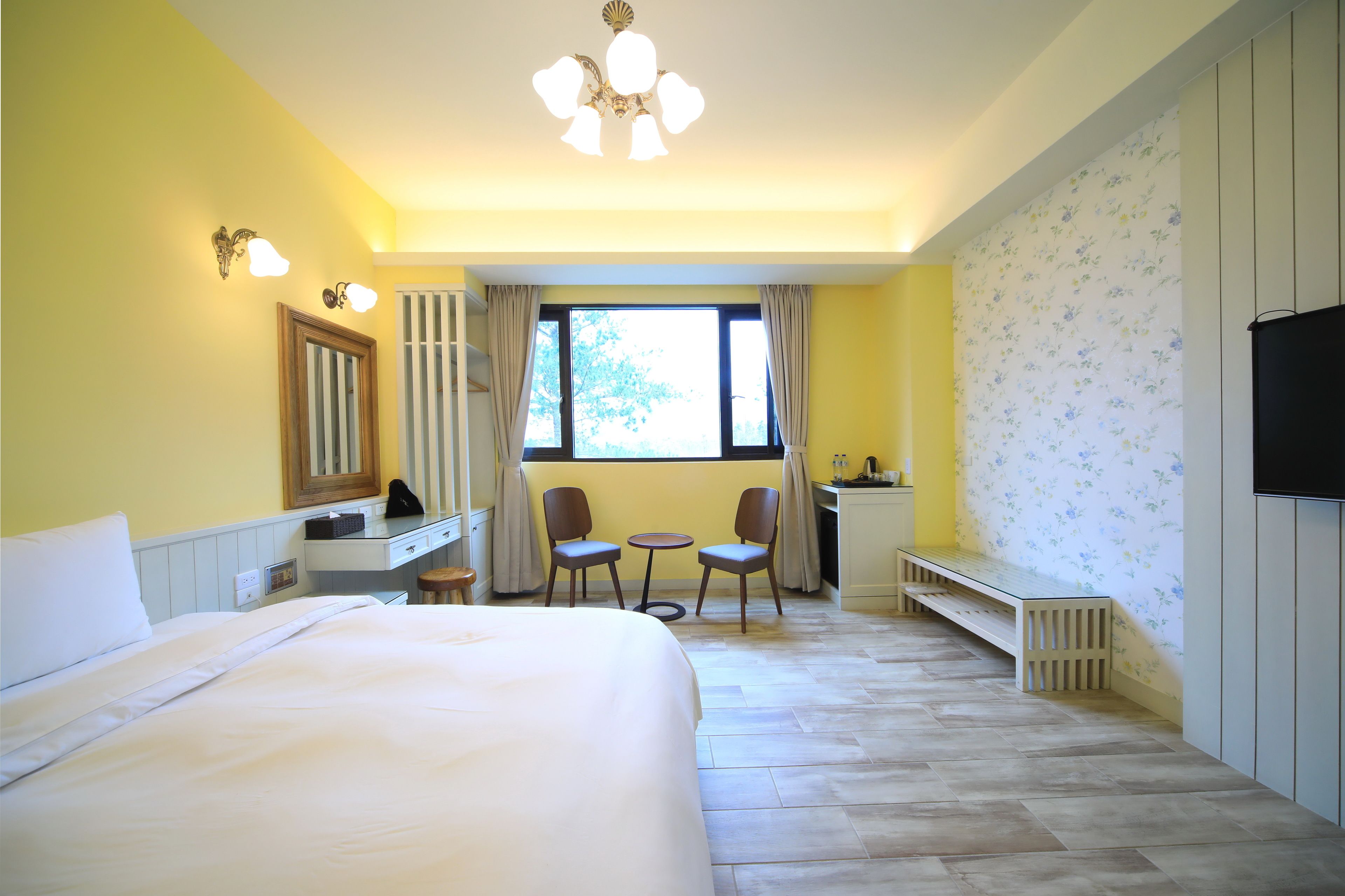 A hotel room with a bed, a desk, two chairs, a small table, a TV, and a window with curtains.