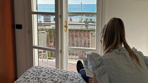 A room with a bed, a window with a view of the sea, and a person reading a book. - Giada Residence (Moneglia)