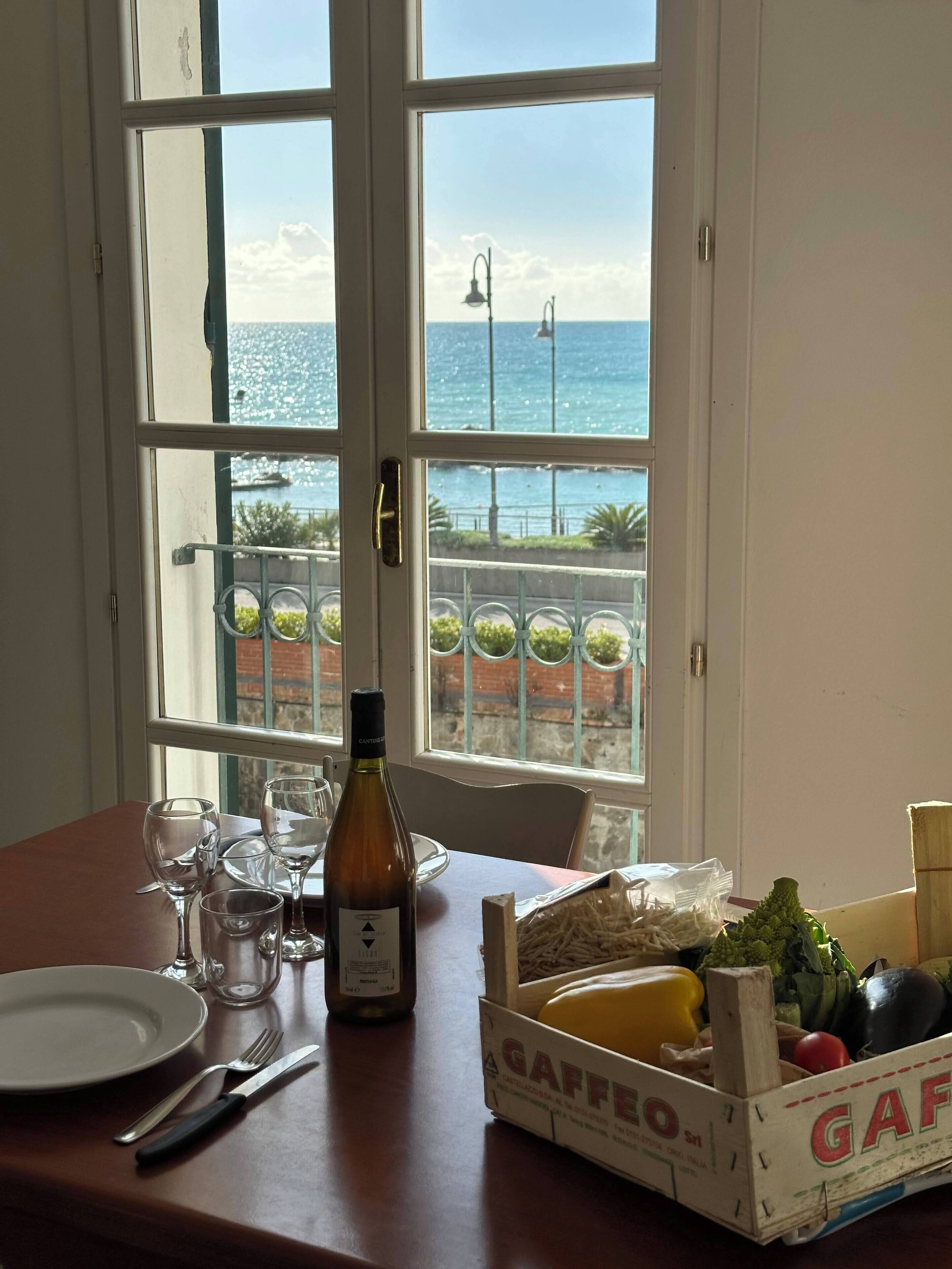 Uma mesa com uma garrafa de vinho, copos, prato, talheres e uma caixa com produtos frescos.