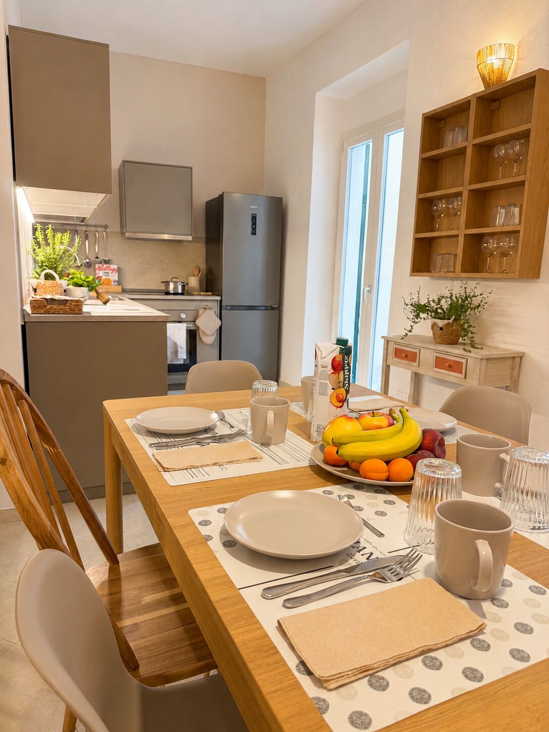 A modern kitchen with a refrigerator, stove, and cabinets. There's a dining table with plates, cutlery, and a fruit bowl.