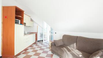 Eine moderne Küche mit weißen Schränken und einem Schachbrettfußboden, ein braunes Sofa und eine Glastür zum Balkon.