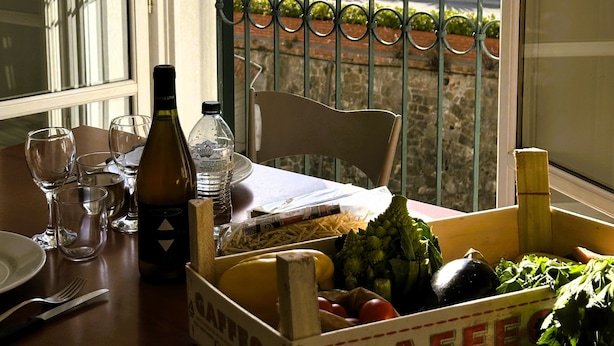 A table with a bottle, glasses, and a crate labeled "GAFFE" containing vegetables, a bottle of water, and a newspaper. - Giada Residence (Moneglia)