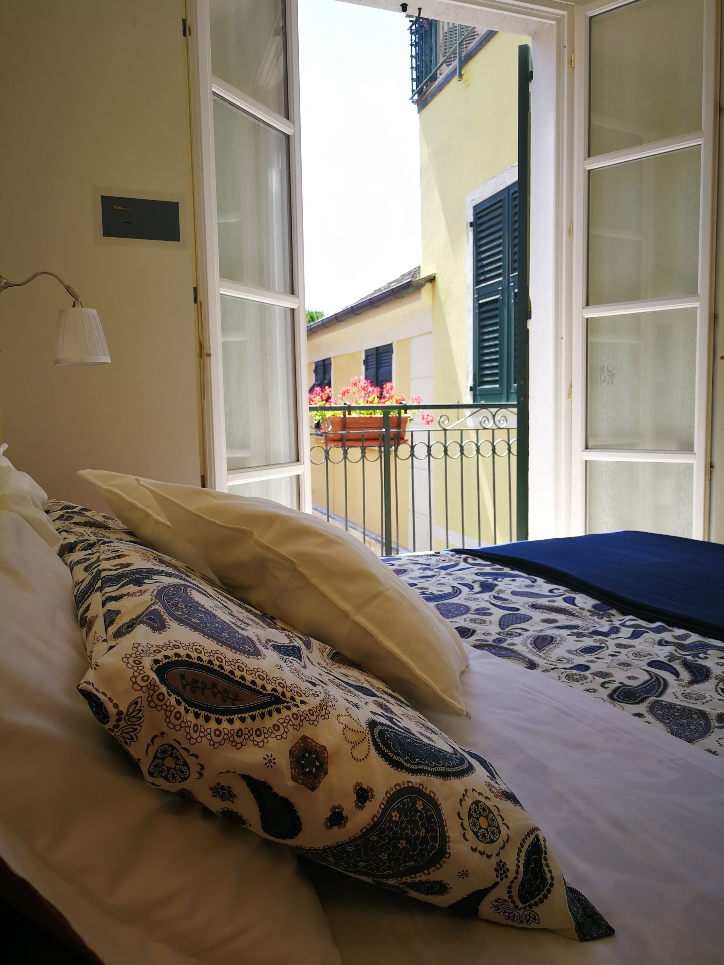 A bed with patterned pillows, a bedside lamp, and a view of a balcony with flowers and a building in the background.