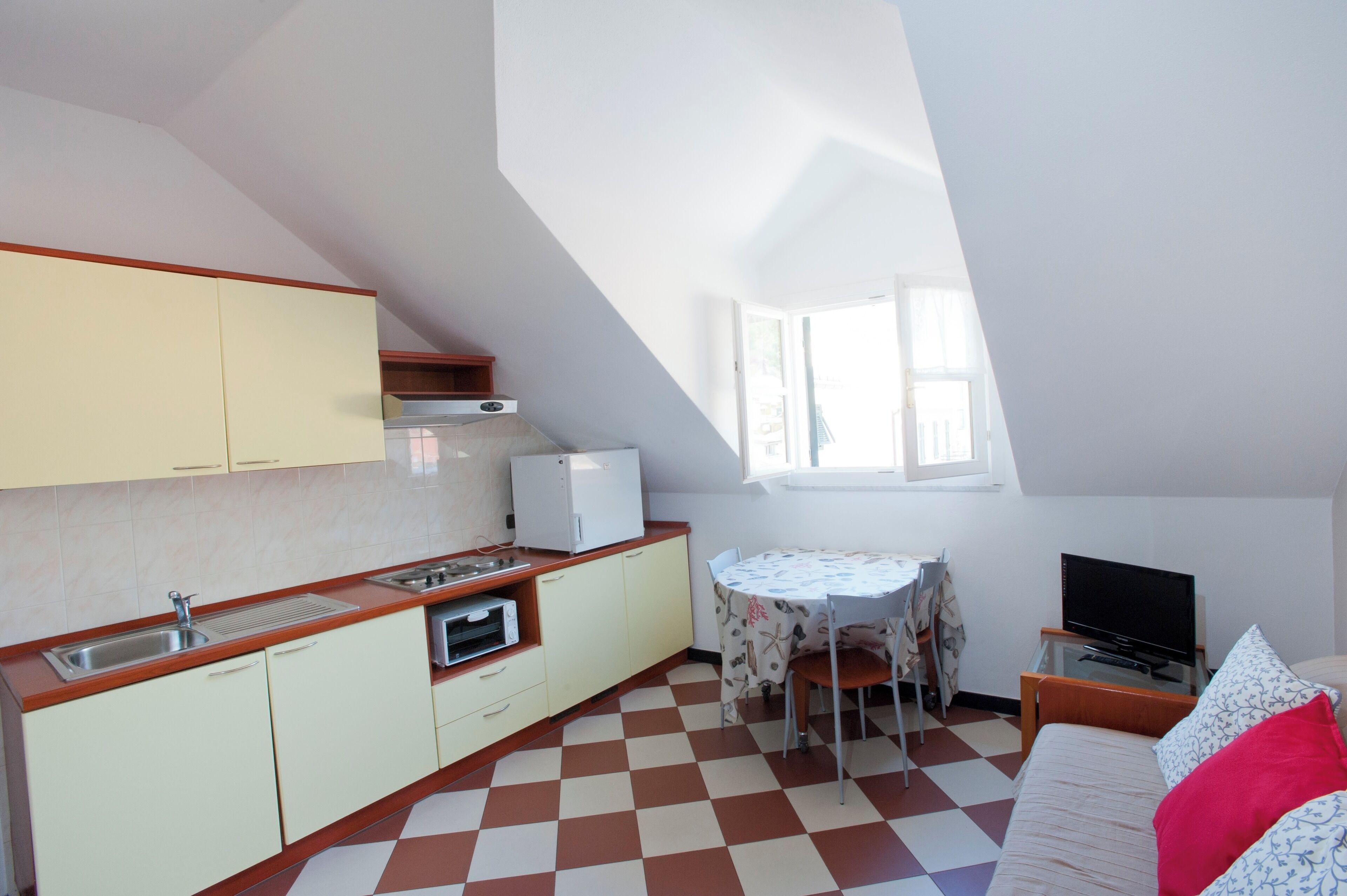 A compact kitchen with a sink, stove, and microwave, a dining table with chairs, a bed with a TV, and a window with natural light.