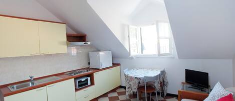 A compact kitchen with a sink, stove, and microwave, a dining table with chairs, a bed with a TV, and a window with natural light.