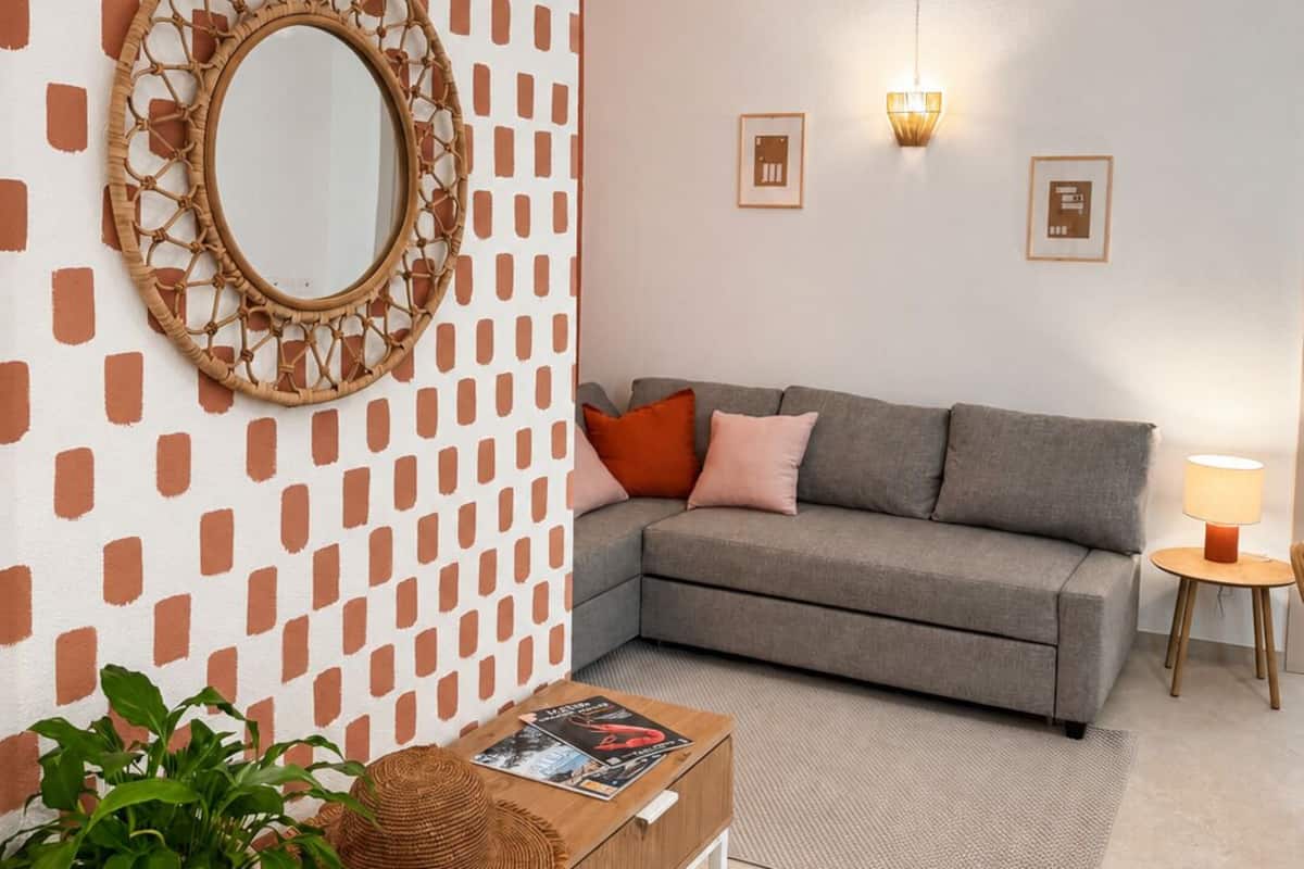 A modern living room with a patterned wall, a round mirror, a grey sofa, a small table with a lamp, and a plant.